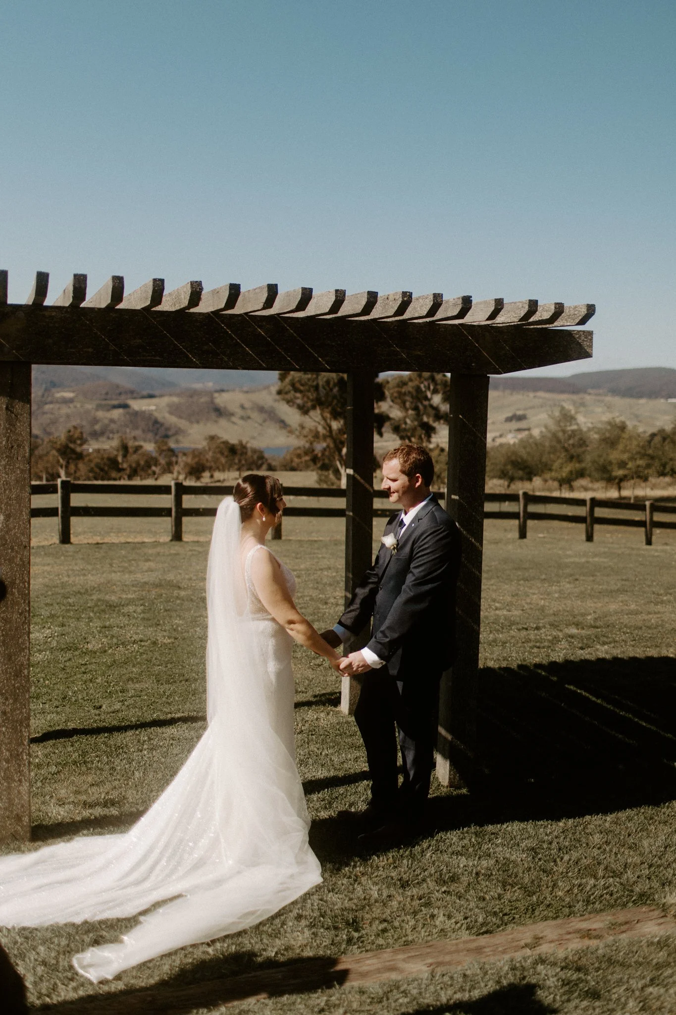 A wedding ceremony taking place outdoors with a bride and groom under a wooden arbor, in a scenic field with trees and mountains in the background at Seclusions Blue Mountains.