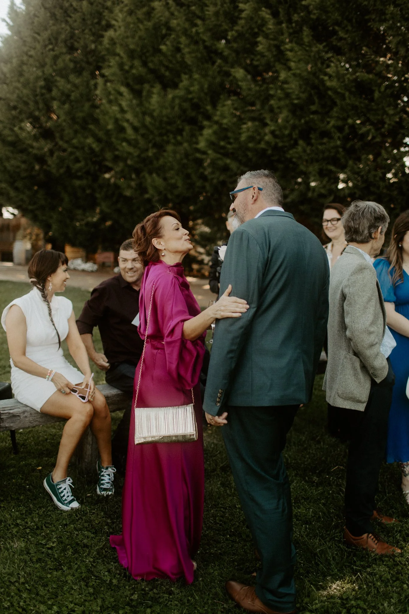 A group of people outdoors during a social gathering, with a woman in a magenta dress and a man in a teal suit engaged in conversation in the foreground, and others sitting and standing nearby.