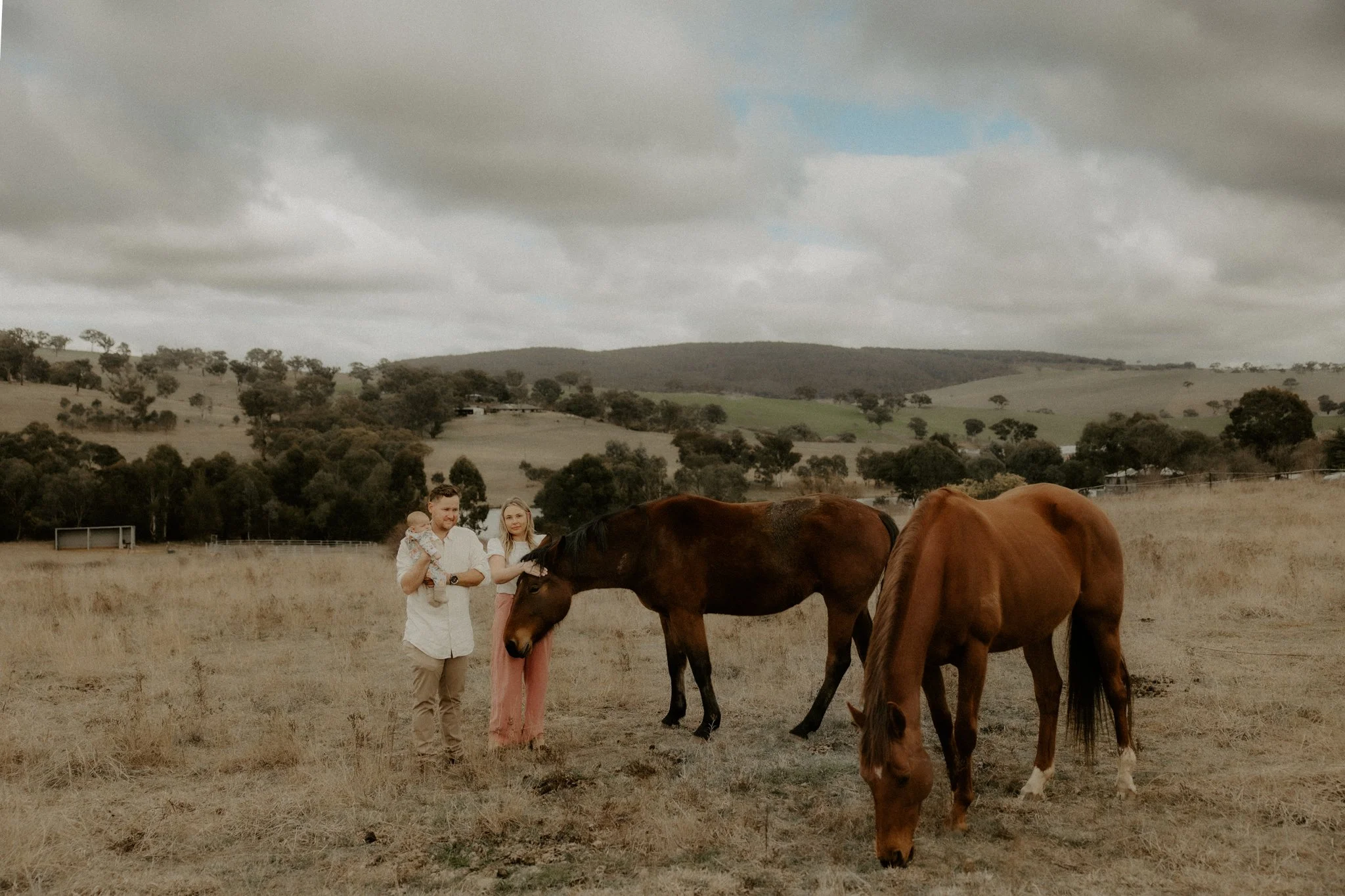 A family of three standing in a field with two horses, one brown and one chestnut, under gray clouds with rolling hills and trees in the background.