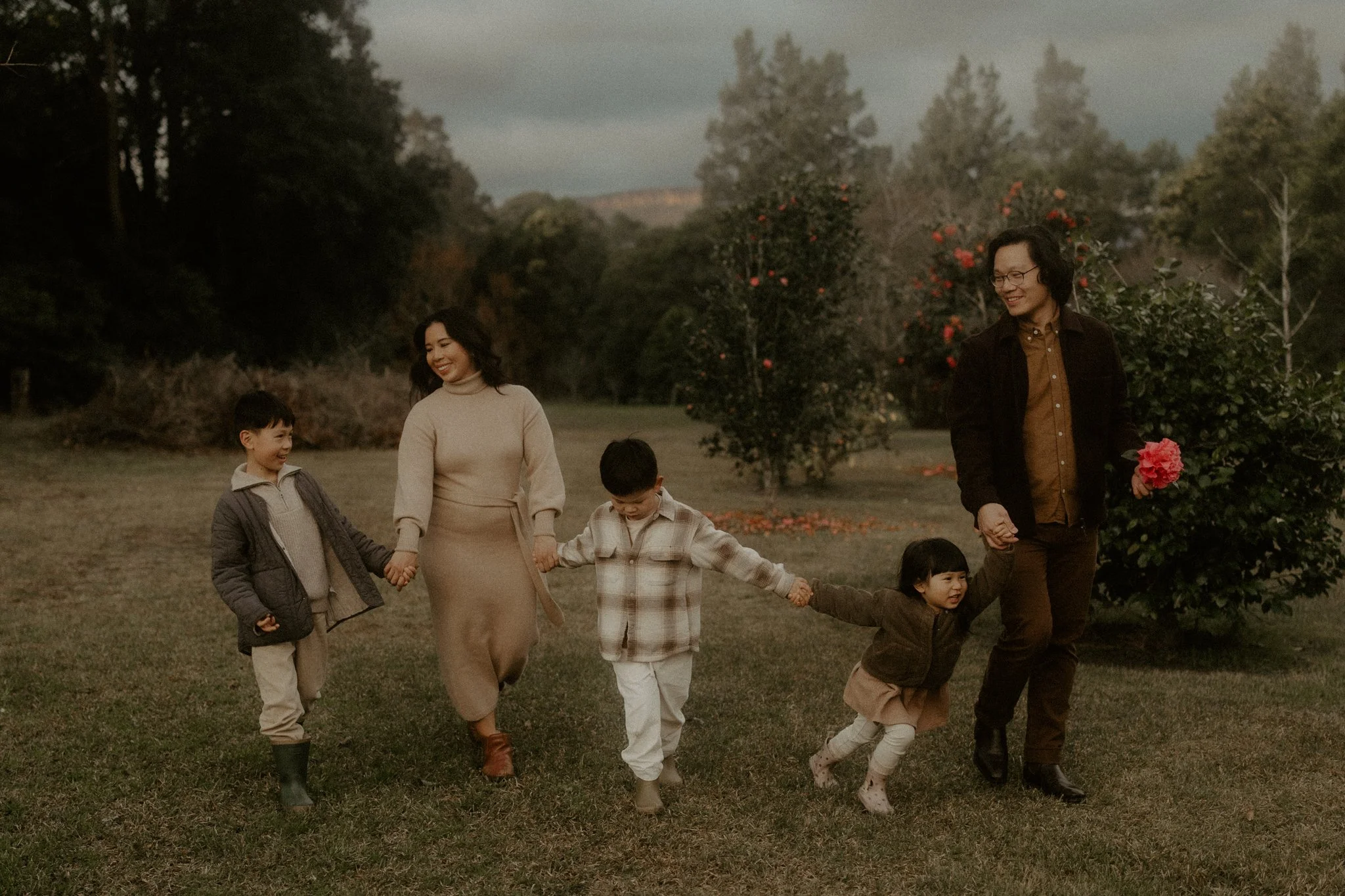 Family of six walking hand in hand in a park, with trees and orange flowers in the background.