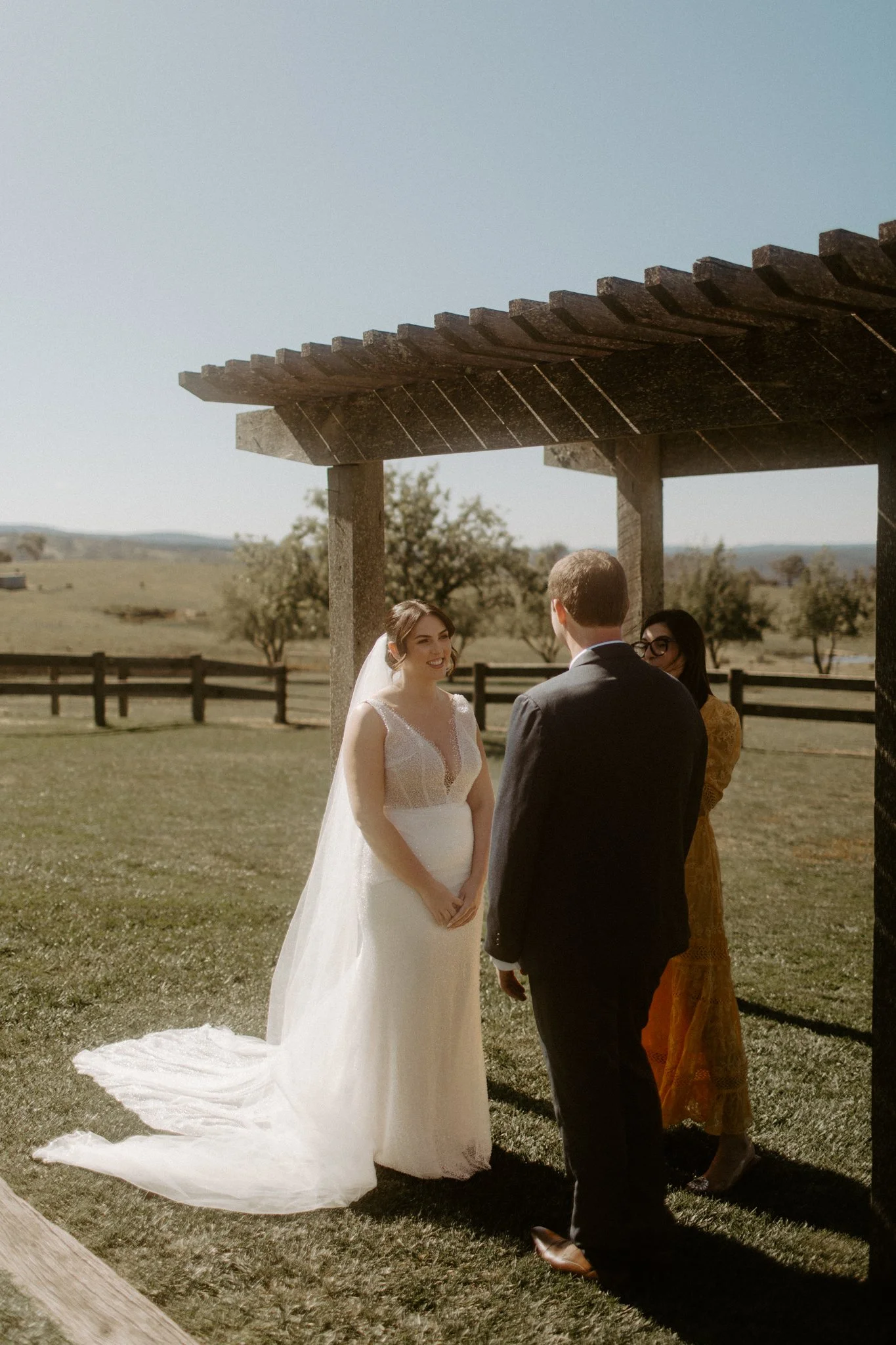 A wedding ceremony taking place outdoors with a bride and groom under a wooden arbor, in a scenic field with trees and mountains in the background at Seclusions Blue Mountains.