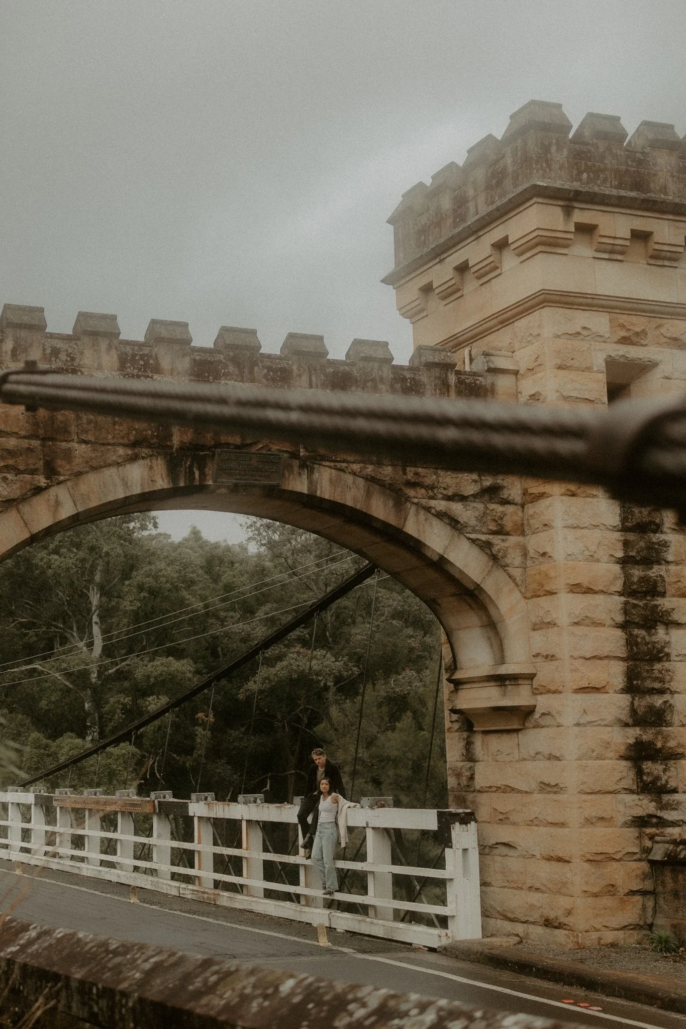 Two people standing on a bridge with an arched stone structure and a castle-like tower behind them, with trees and an overcast sky in the background.