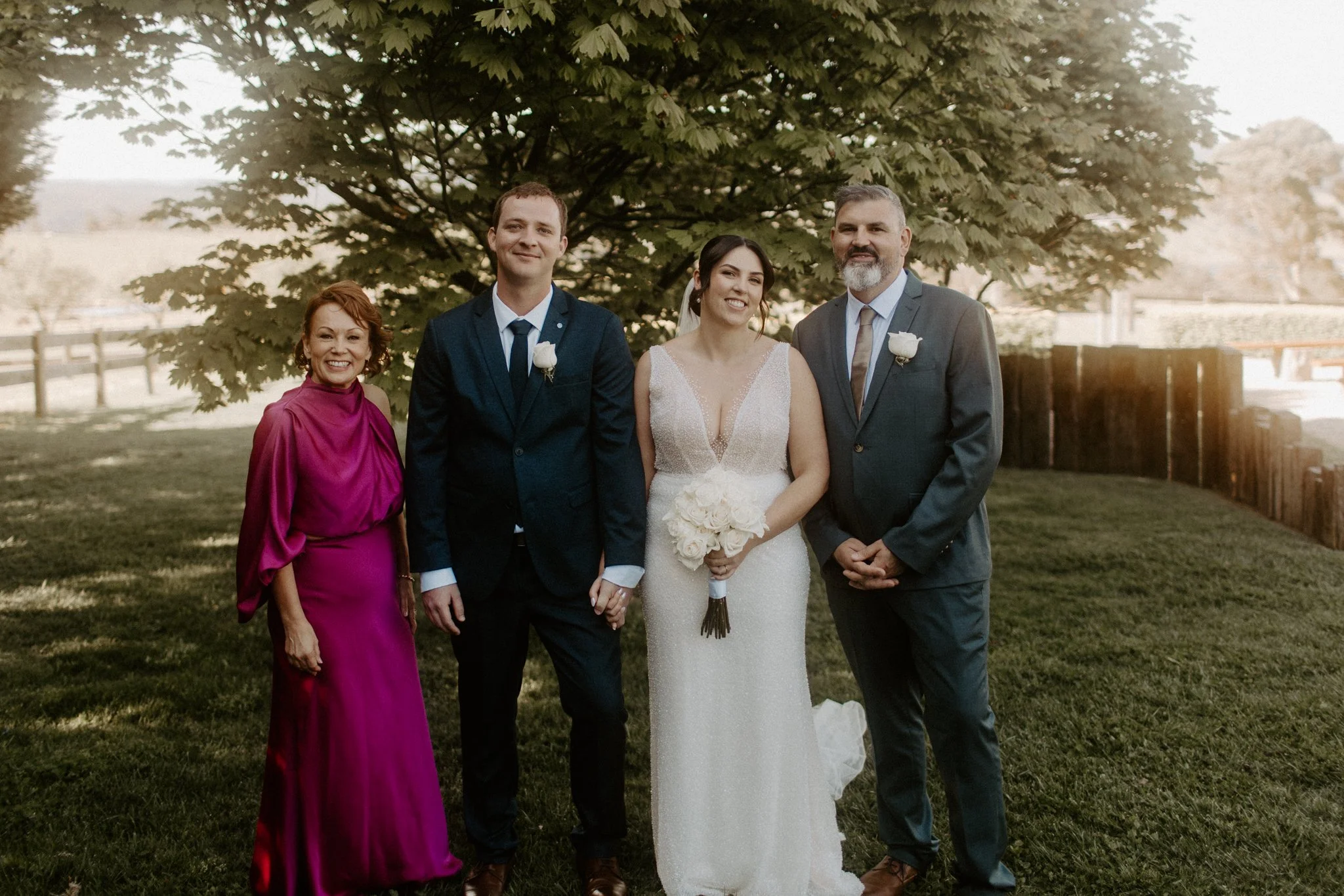 A wedding photo taken outdoors featuring four people standing on grass beneath a large leafy tree. From left to right: a woman in a magenta gown with short red hair, a man in a dark blue suit with a white shirt and tie, a bride in a white wedding gow