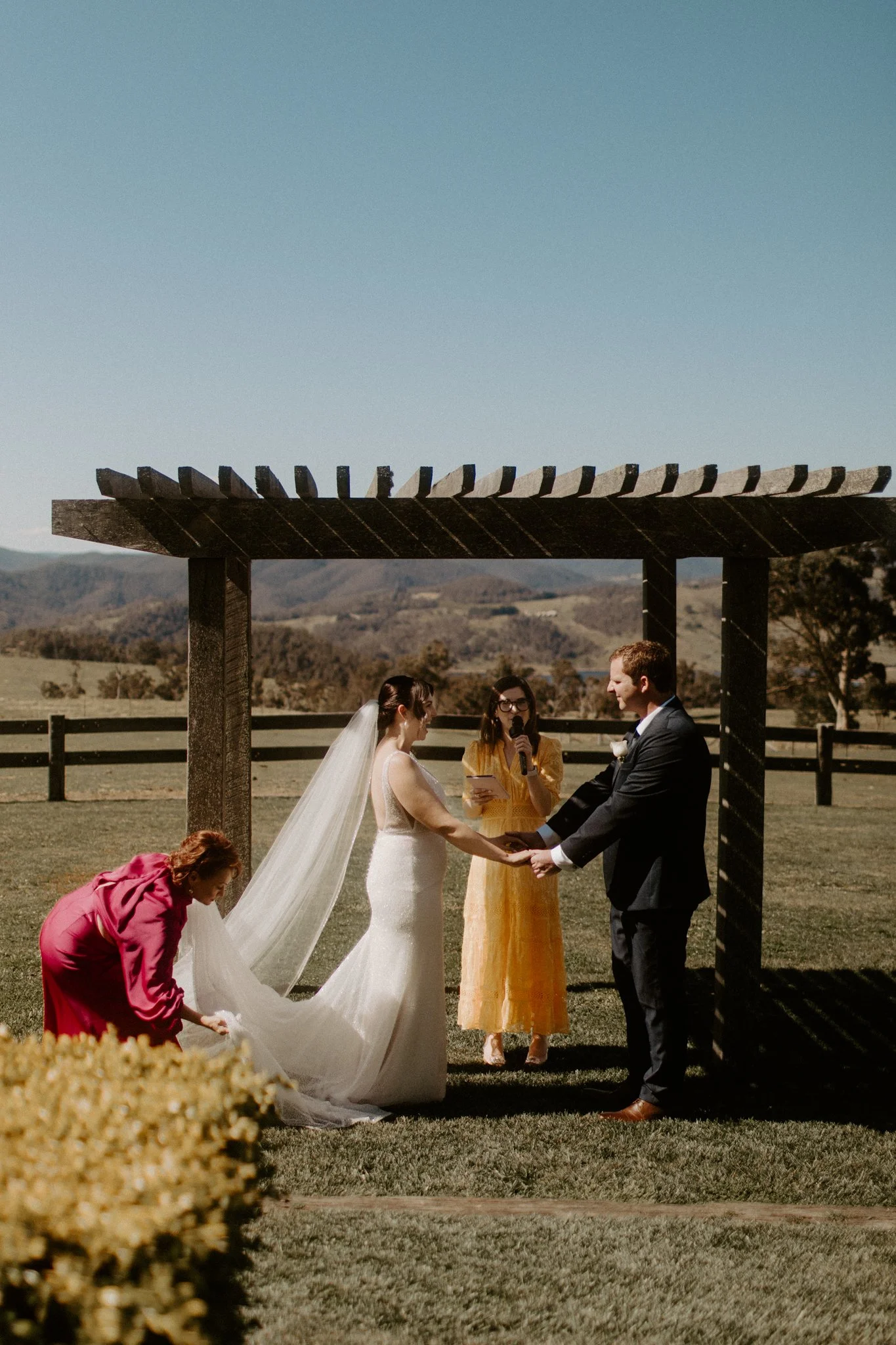 A wedding ceremony taking place outdoors with a bride and groom facing officiant under a wooden arbor, in a scenic field with trees and mountains in the background at Seclusions Blue Mountains.
