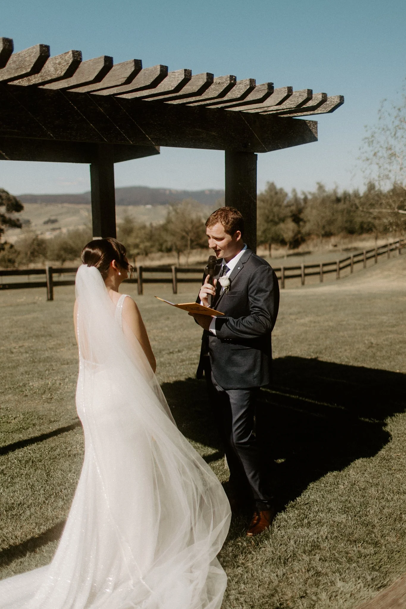 A wedding ceremony taking place outdoors with a bride and groom under a wooden arbor, in a scenic field with trees and mountains in the background at Seclusions Blue Mountains.