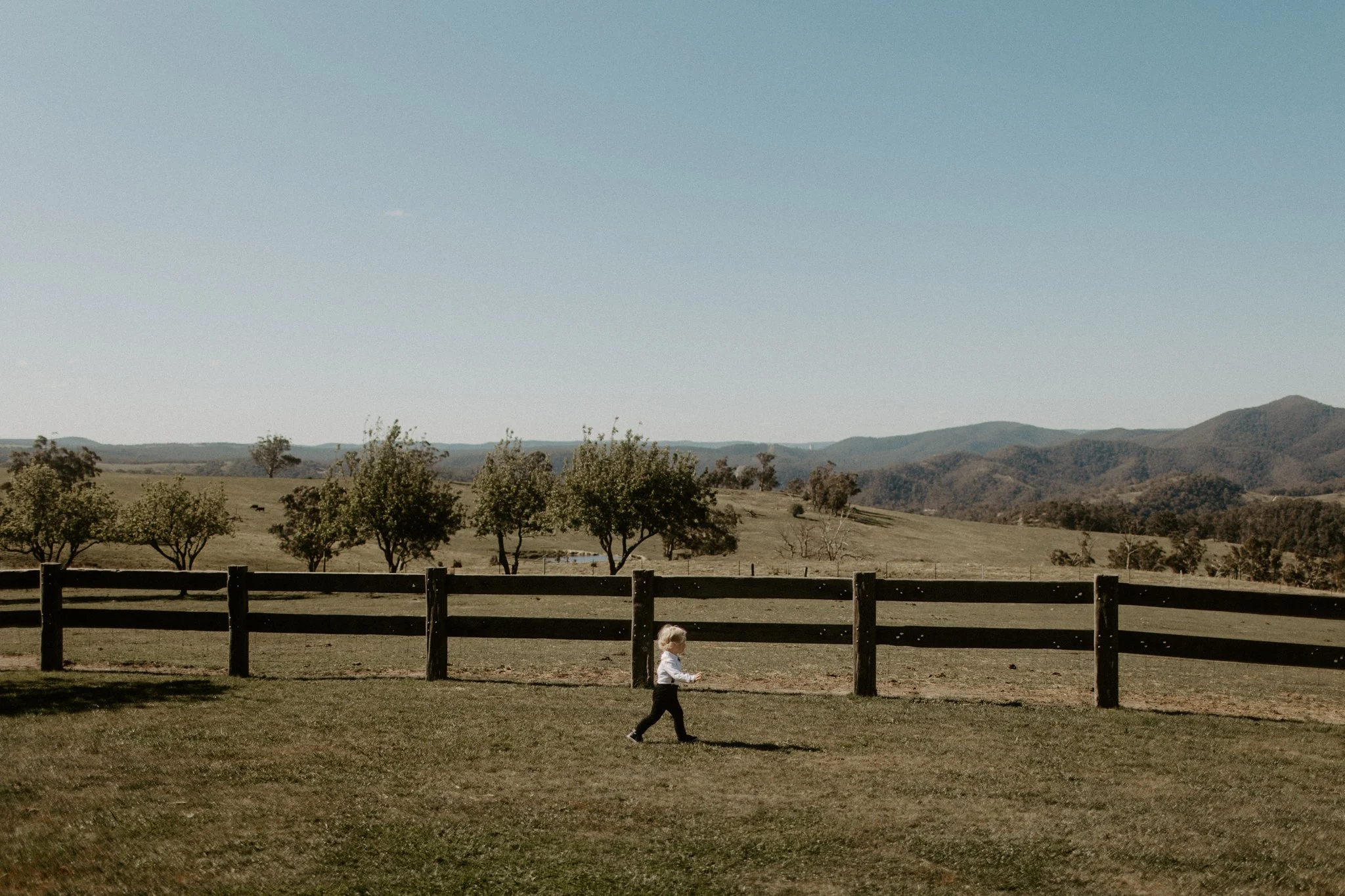A young child running on a grassy lawn in front of a wooden fence. The background features rolling hills with scattered trees and a clear blue sky during a Blue Mountains wedding at Seclusions Blue Mountains.