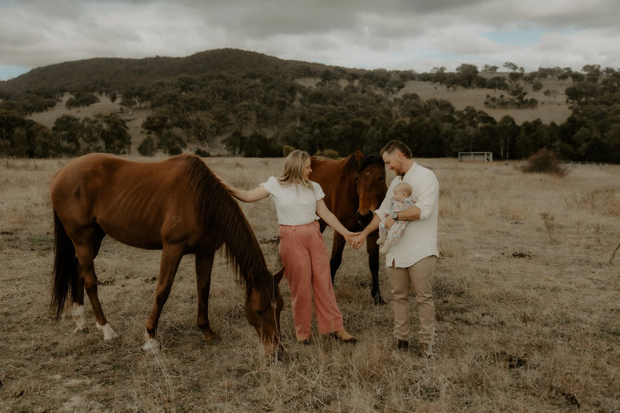 A family of four standing in a grassy field with two brown horses. The woman is holding one horse's head, the man is holding a baby, and the woman is touching the other horse.