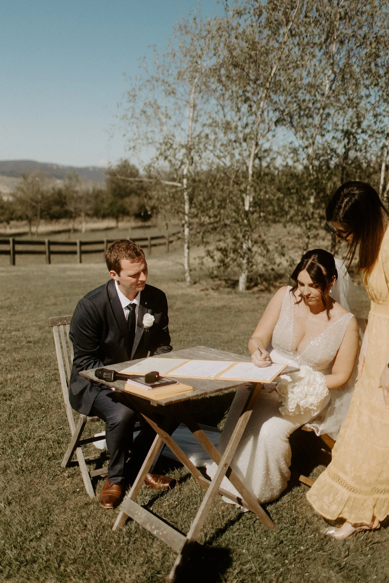 A bride and groom sitting outdoors during a wedding ceremony, with a woman standing and signing a document during a Blue Mountains wedding at Seclusions Blue Mountains.