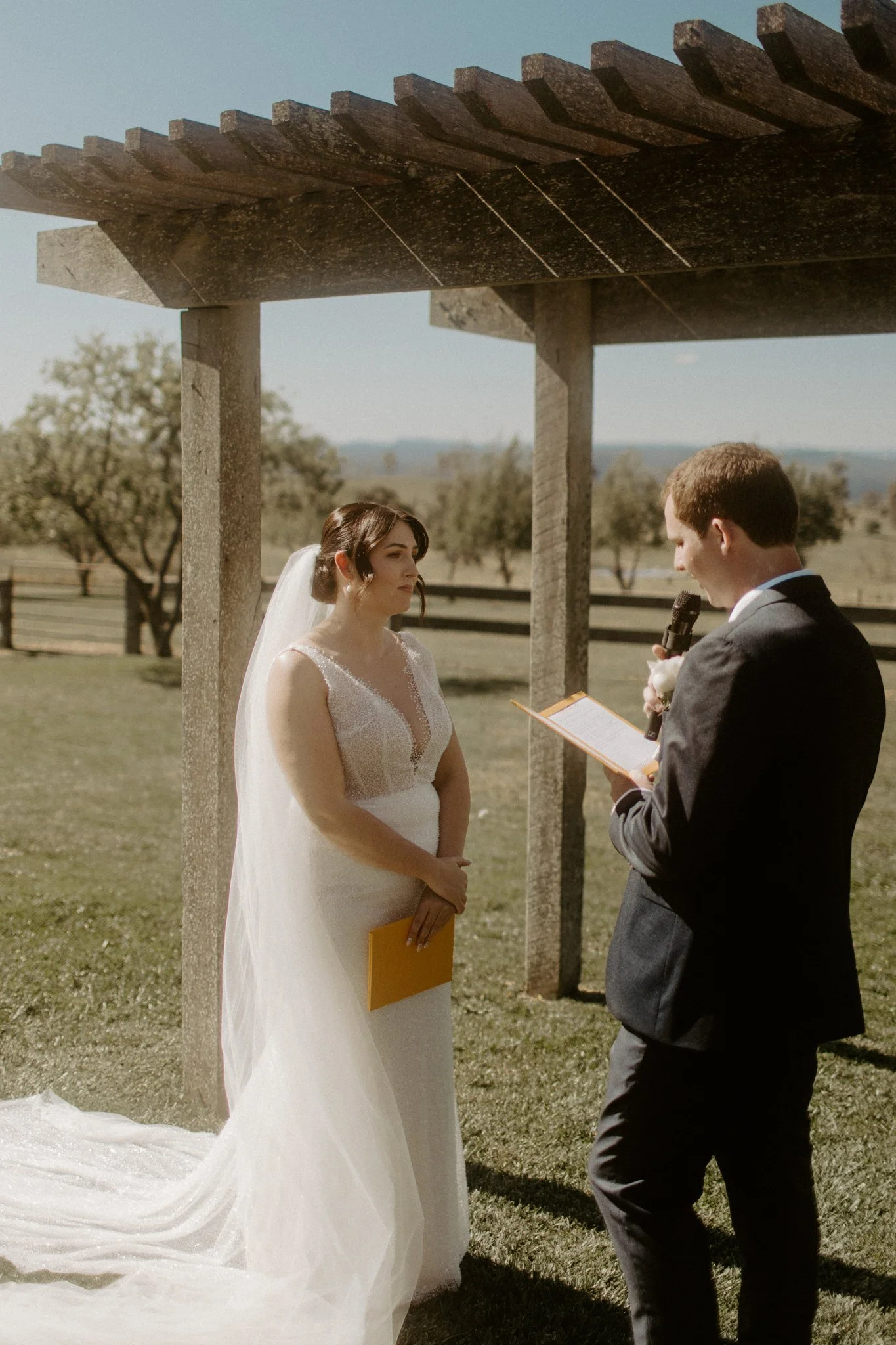 A wedding ceremony taking place outdoors with a bride and groom under a wooden arbor, in a scenic field with trees and mountains in the background at Seclusions Blue Mountains.