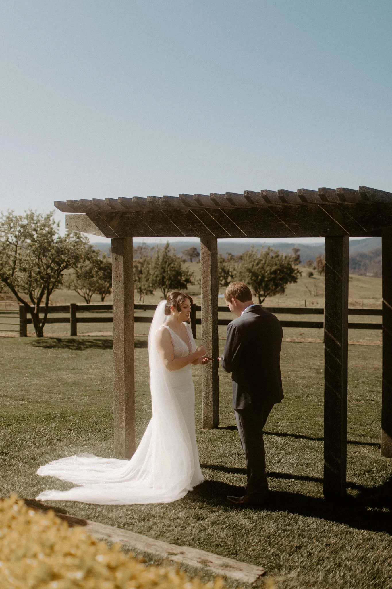 A wedding ceremony taking place outdoors with a bride and groom under a wooden arbor, in a scenic field with trees and mountains in the background at Seclusions Blue Mountains.