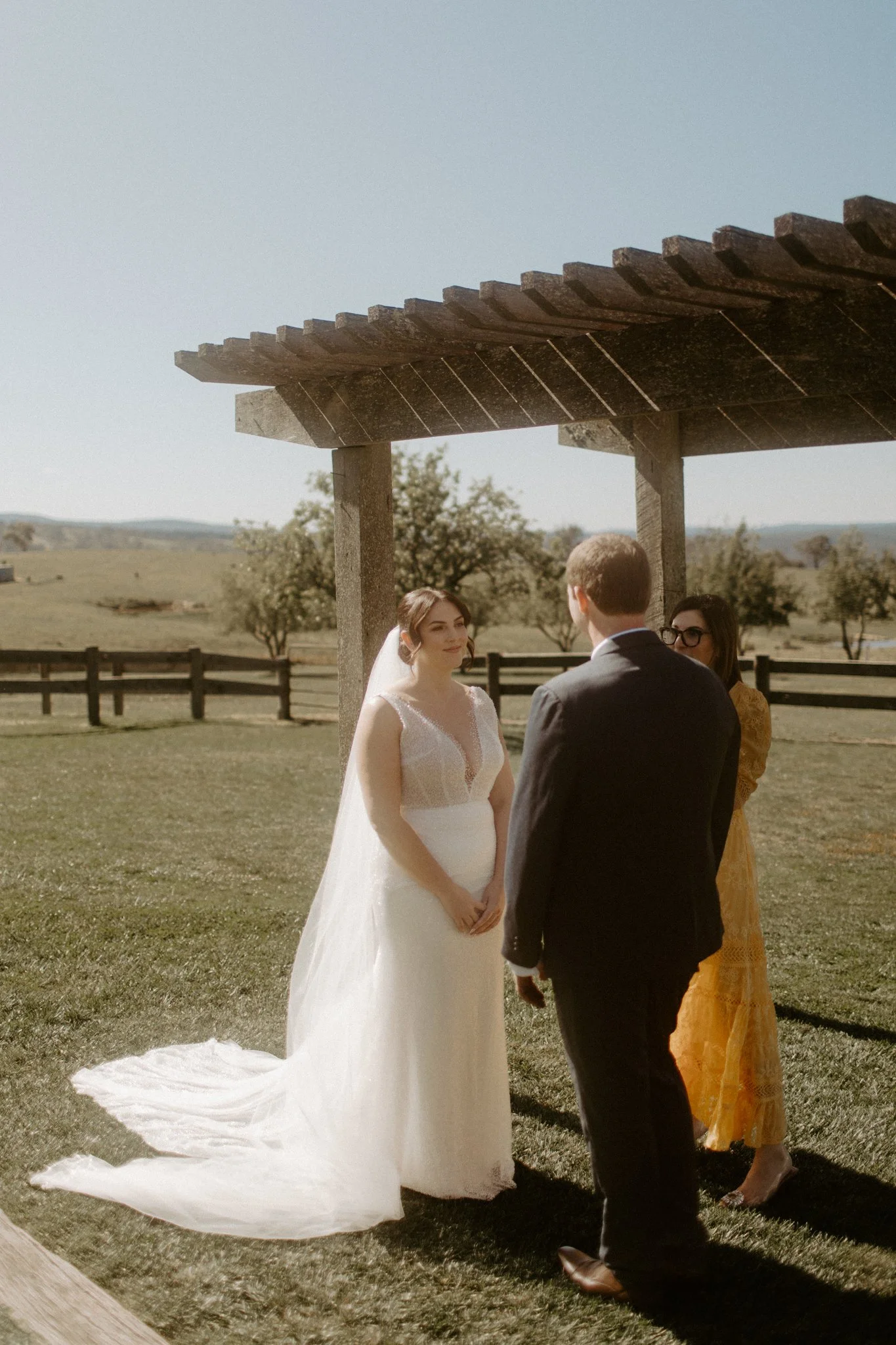 A wedding ceremony taking place outdoors with a bride and groom under a wooden arbor, in a scenic field with trees and mountains in the background at Seclusions Blue Mountains.