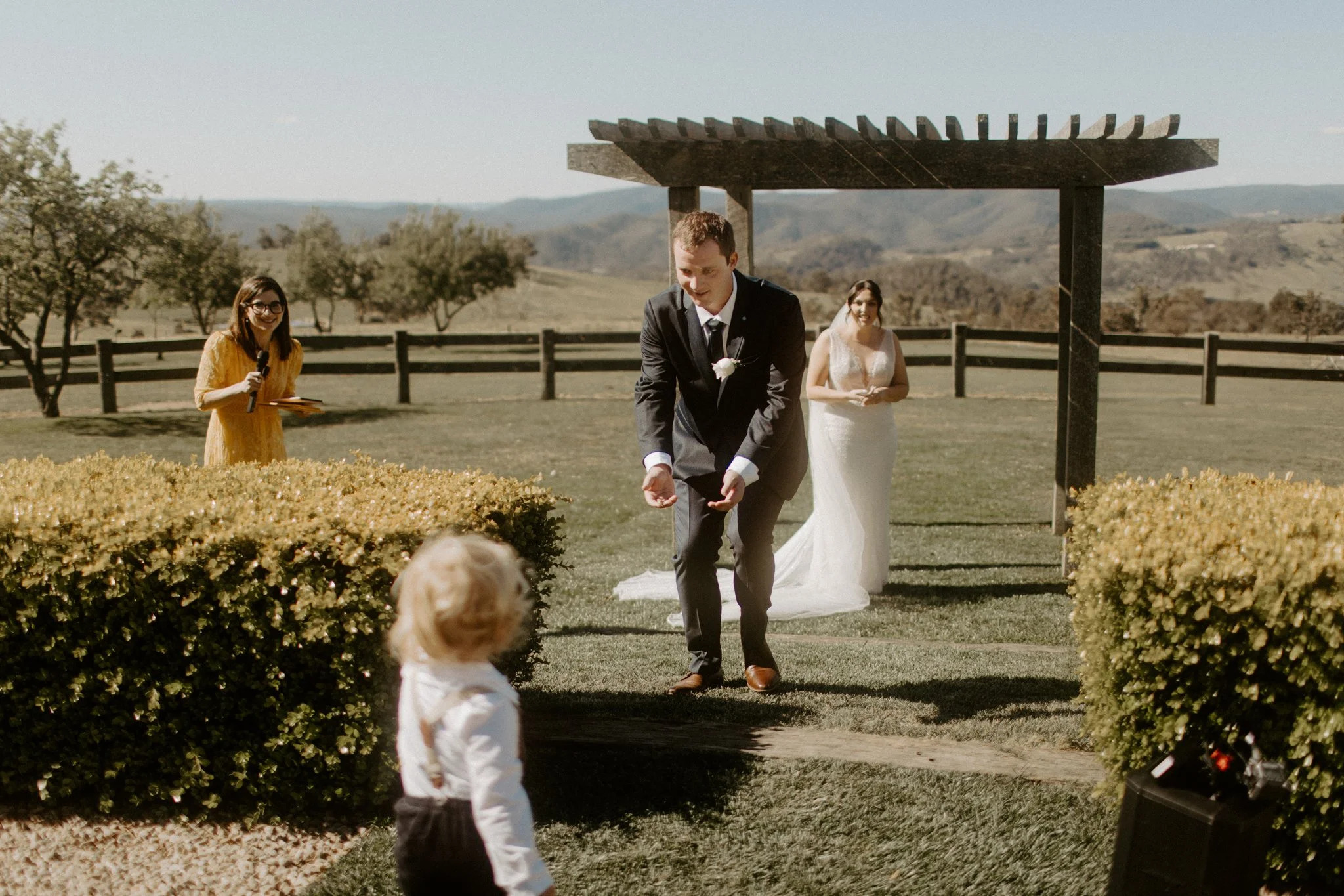 A wedding ceremony outdoors with a groom adjusting his suit, a bride in a white gown behind him, an officiant holding a microphone, and a young child watching, set in a scenic rural landscape.