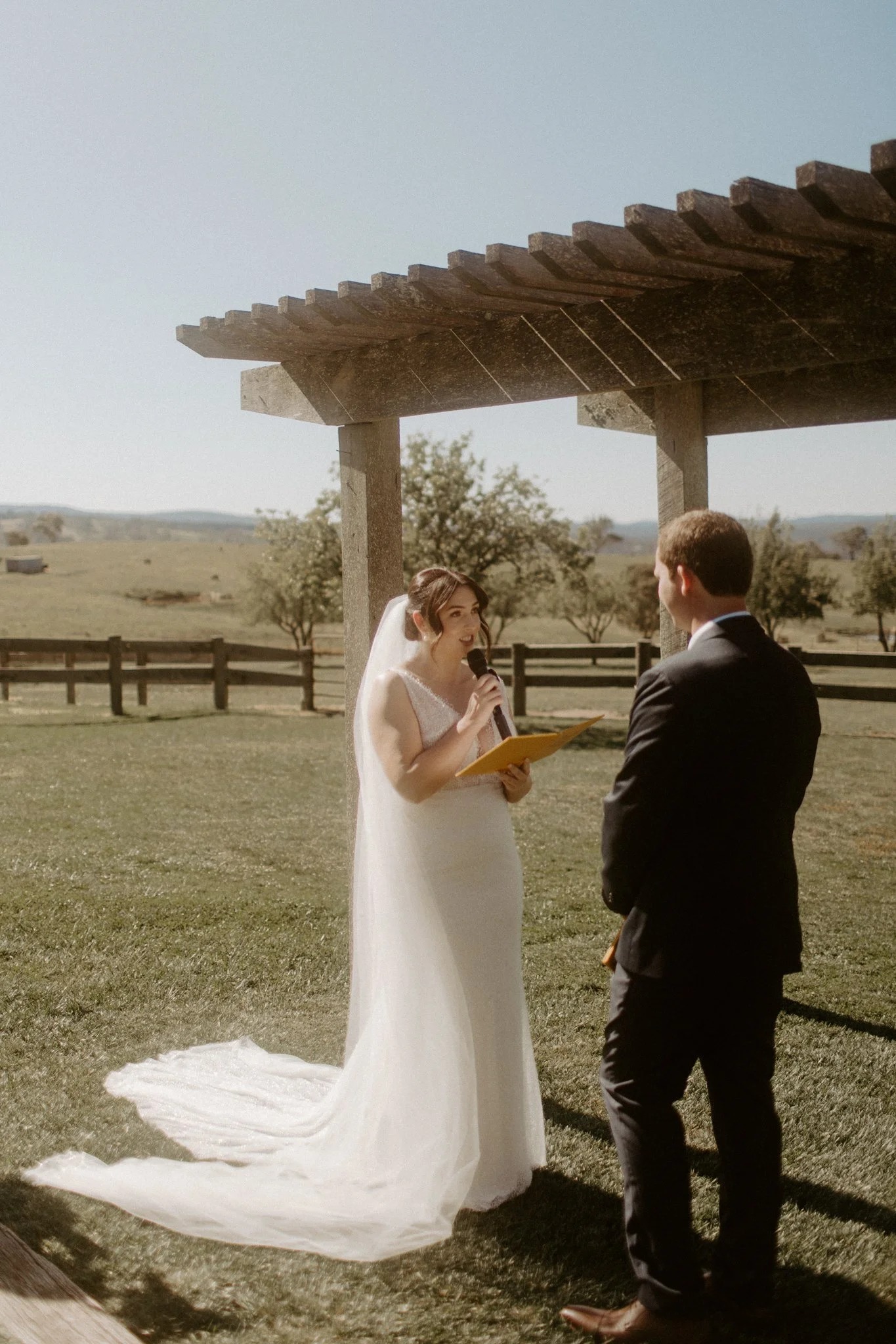 A wedding ceremony taking place outdoors with a bride and groom under a wooden arbor, in a scenic field with trees and mountains in the background at Seclusions Blue Mountains.