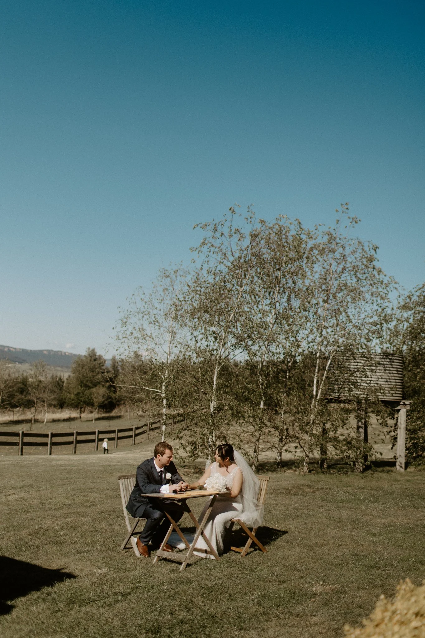 A bride and groom sitting at a small wooden table outdoors, holding hands during a Blue Mountains wedding at Seclusions Blue Mountains.