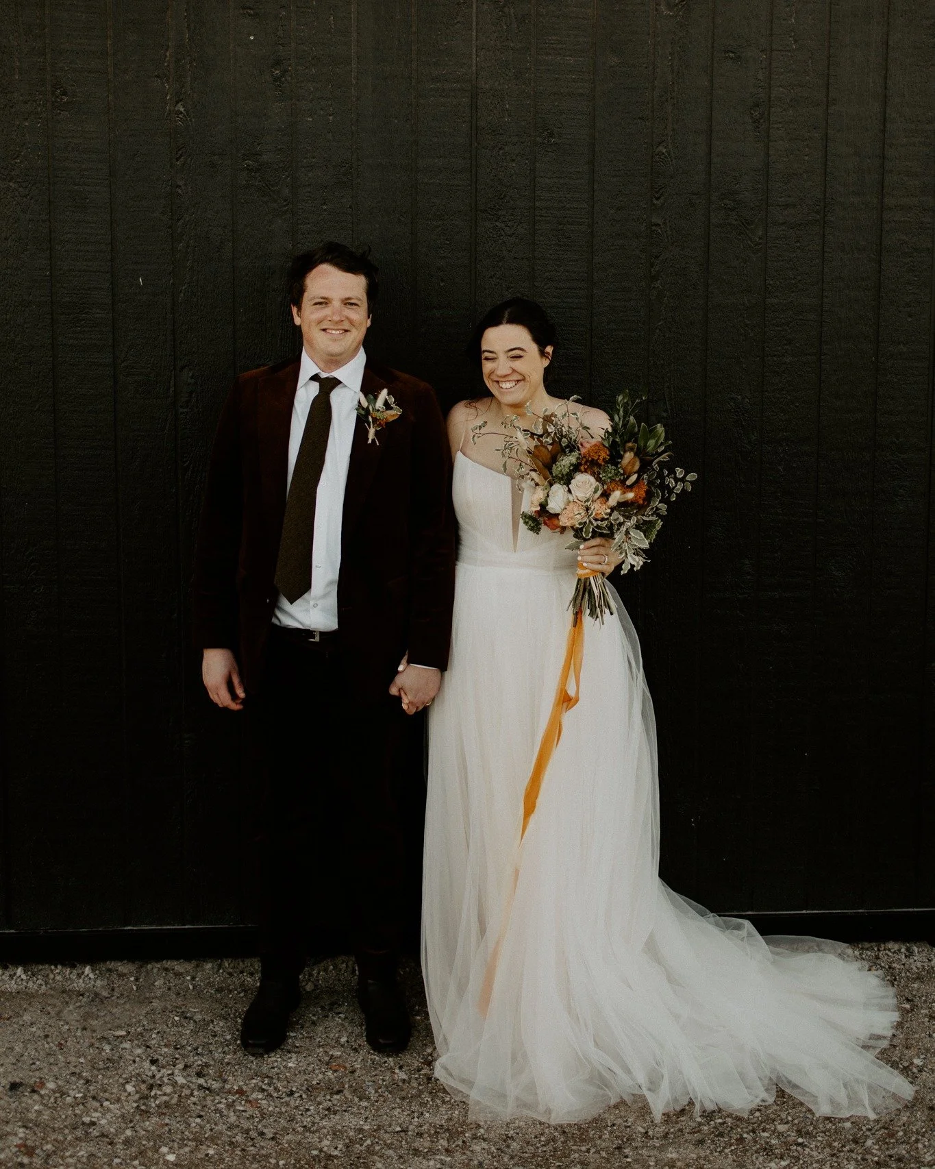 A bride and groom standing hand in hand outdoors, smiling, with a dark wooden wall in the background. The bride wears a white wedding dress and holds a colorful bouquet, while the groom wears a dark suit with a white shirt and dark tie.
