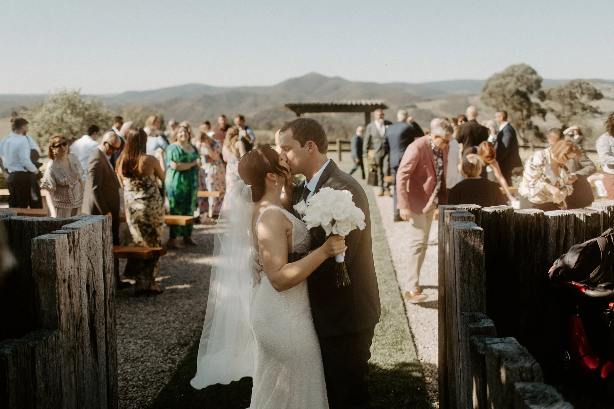 A bride and groom sharing a kiss at their outdoor wedding, with guests standing and seated in the background amidst a scenic landscape during a Blue Mountains wedding at Seclusions Blue Mountains.