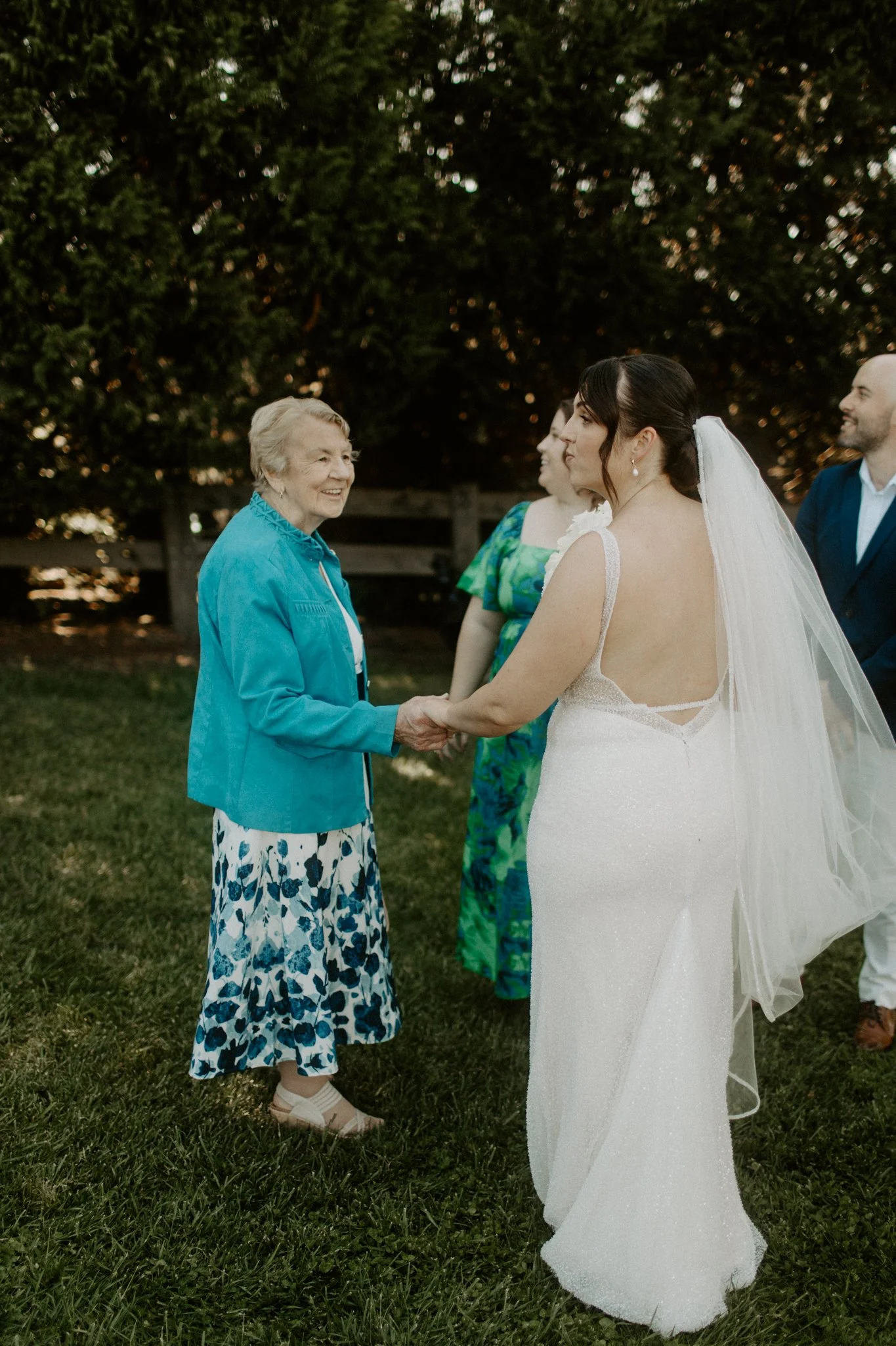 A bride in a white wedding dress holding hands and smiling at an elderly woman in a turquoise jacket and floral skirt, outdoors during a wedding celebration at sunset.