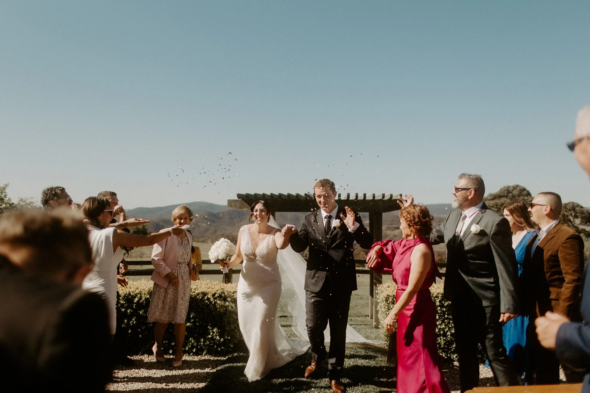 A bride and groom walking down the aisle outdoors with guests on either side, some tossing flower petals in the air during a Blue Mountains wedding at Seclusions Blue Mountains.