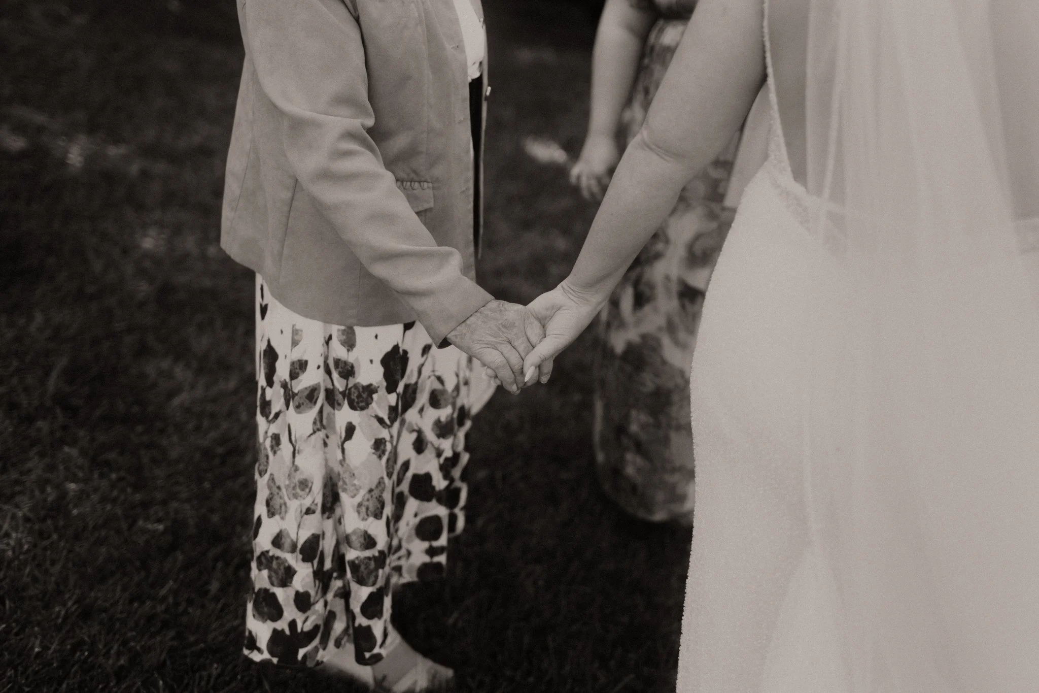 An elderly woman and a bride hold hands during a wedding ceremony outdoors.