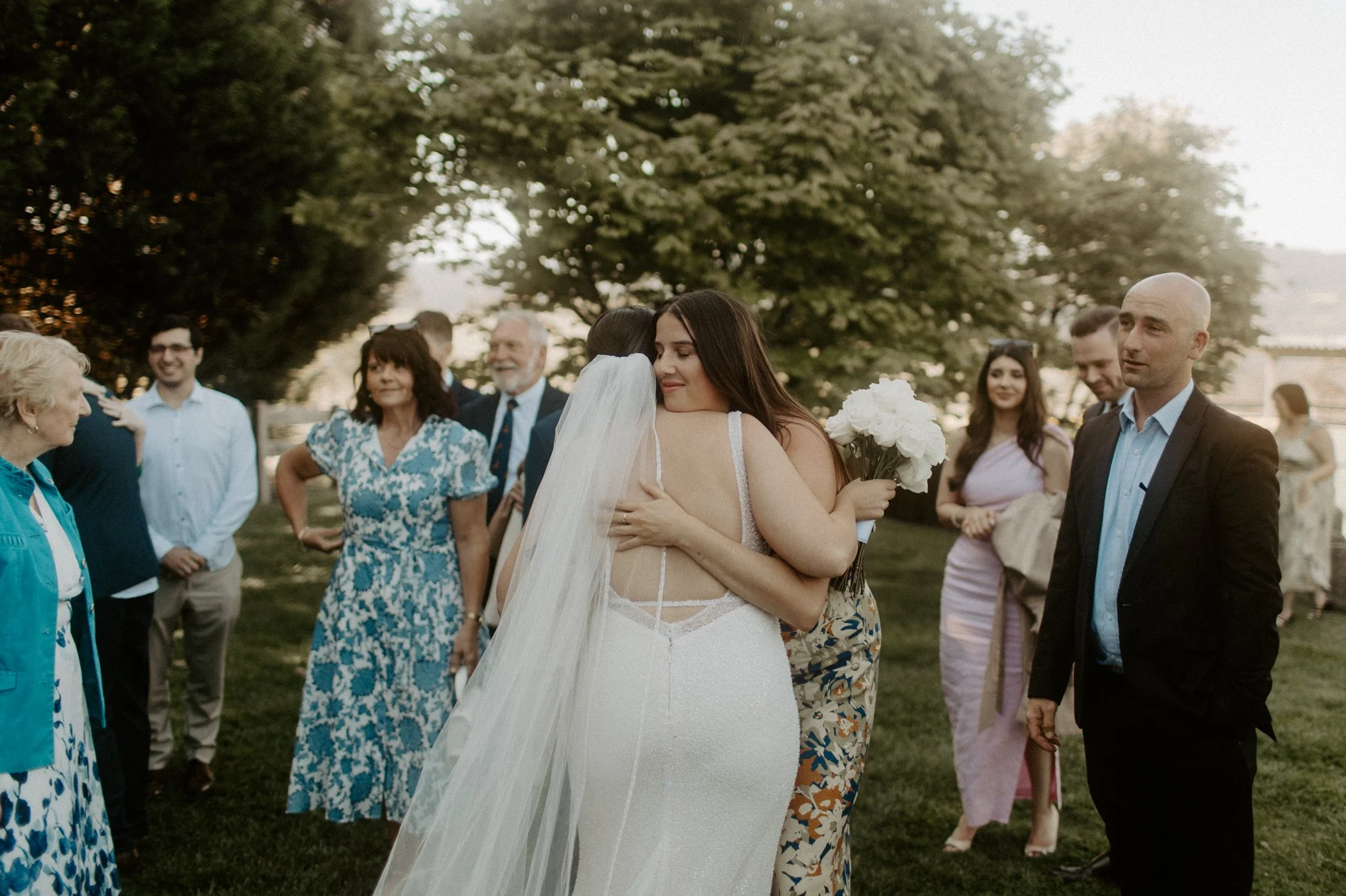 Two women hugging at a wedding reception outdoors, one in a white wedding gown with a veil and the other in a floral dress holding a bouquet of white flowers, with friends and family in the background on a grassy area with trees.