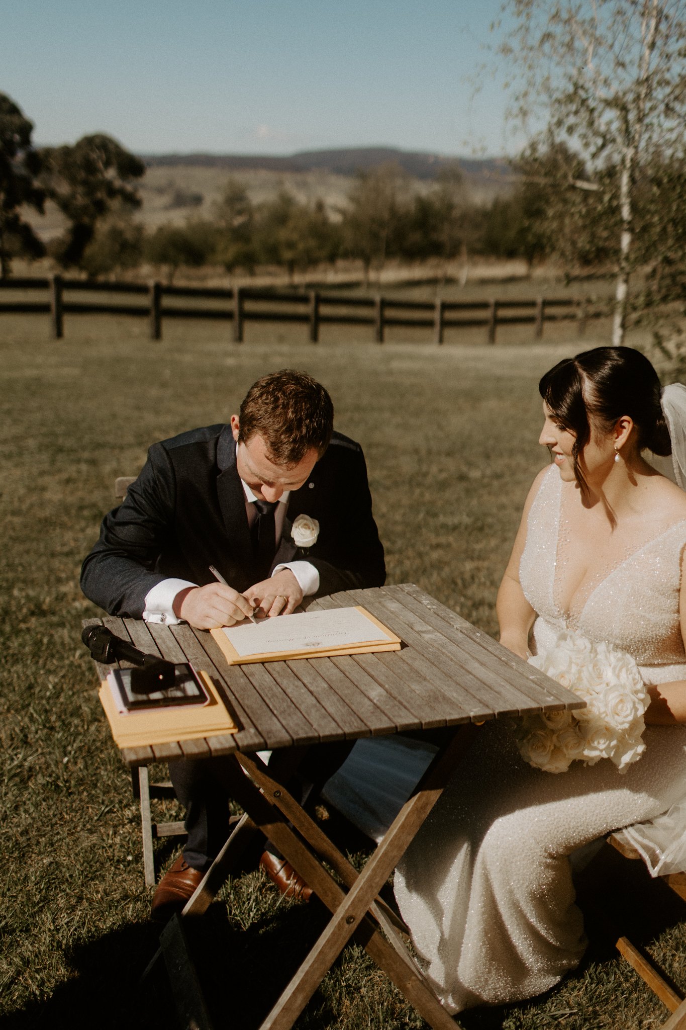 A wedding ceremony outdoors with a bride in a white gown and a groom in a black suit sitting at a wooden table. The groom is signing a document while the bride watches and smiles during a Blue Mountains wedding at Seclusions Blue Mountains.