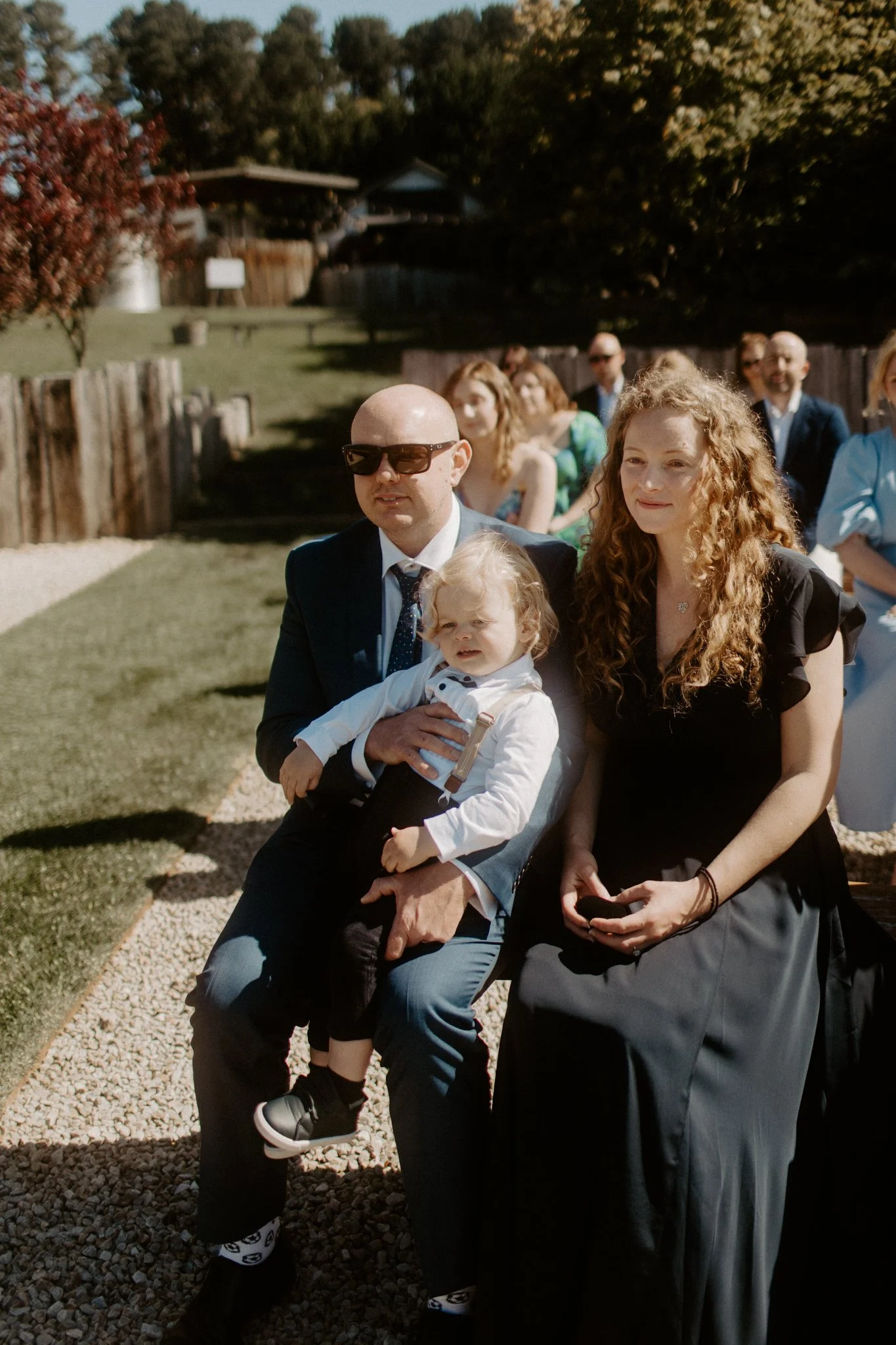 A group of people attending an outdoor wedding ceremony at Seclusions Blue Mountains, sitting on wooden benches, with trees and a blue sky in the background.
