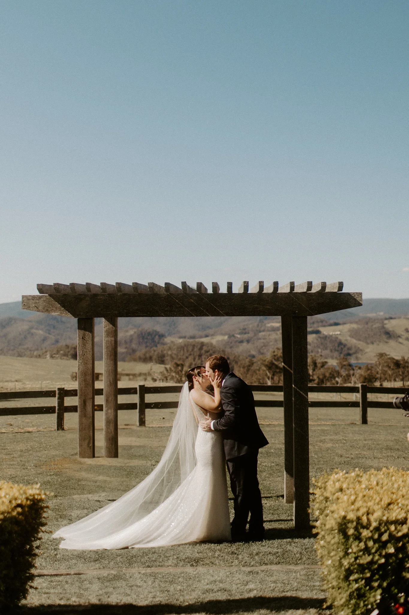 A bride and groom sharing a kiss outdoors under a wooden structure with mountains in the background during a Blue Mountains wedding at Seclusions Blue Mountains.