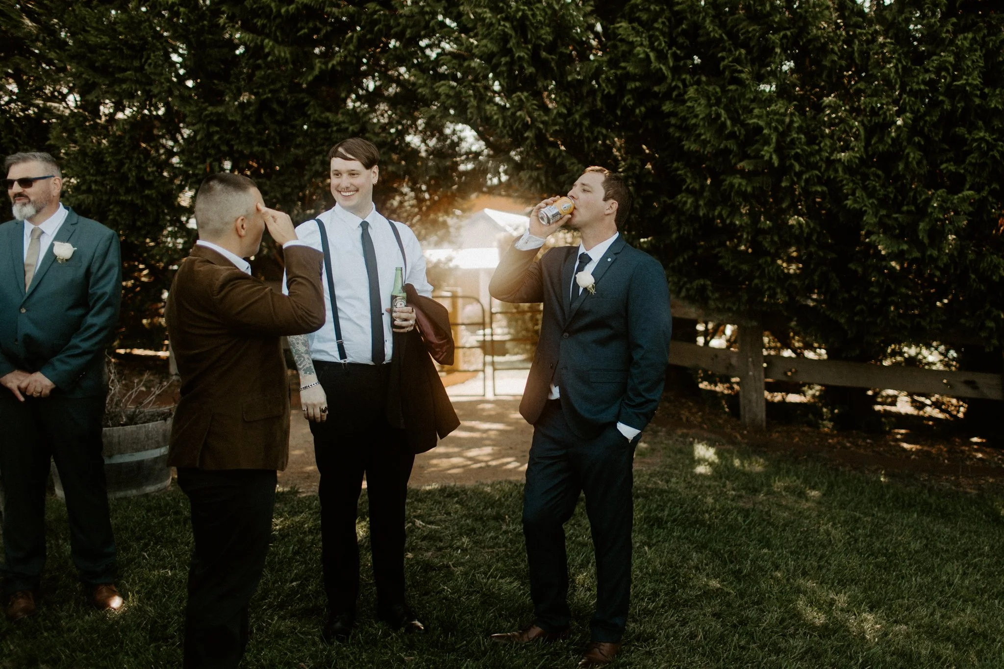 Group of men dressed in suits at outdoor event, some holding drinks, standing on grass with trees and wooden fence in background.