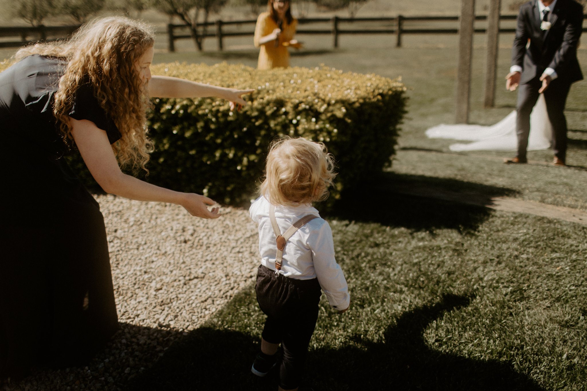 A woman with long curly hair pointing at a young child dressed in formal attire, walking outside near bushes and a gravel path, with a bride and groom in the background.