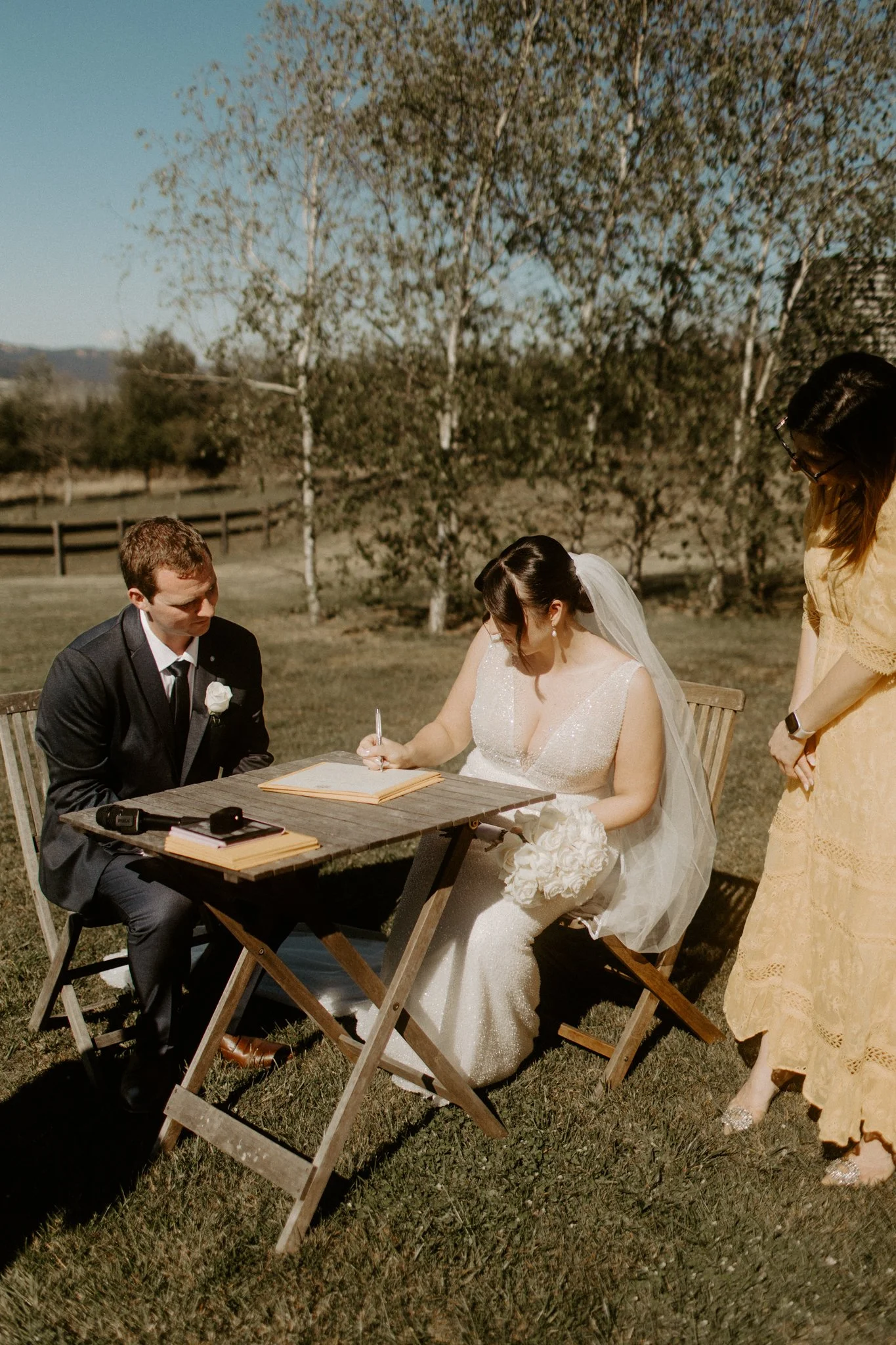 A bride and groom seated outdoors at a wedding ceremony, signing a document, with a woman standing beside them during a Blue Mountains wedding at Seclusions Blue Mountains.