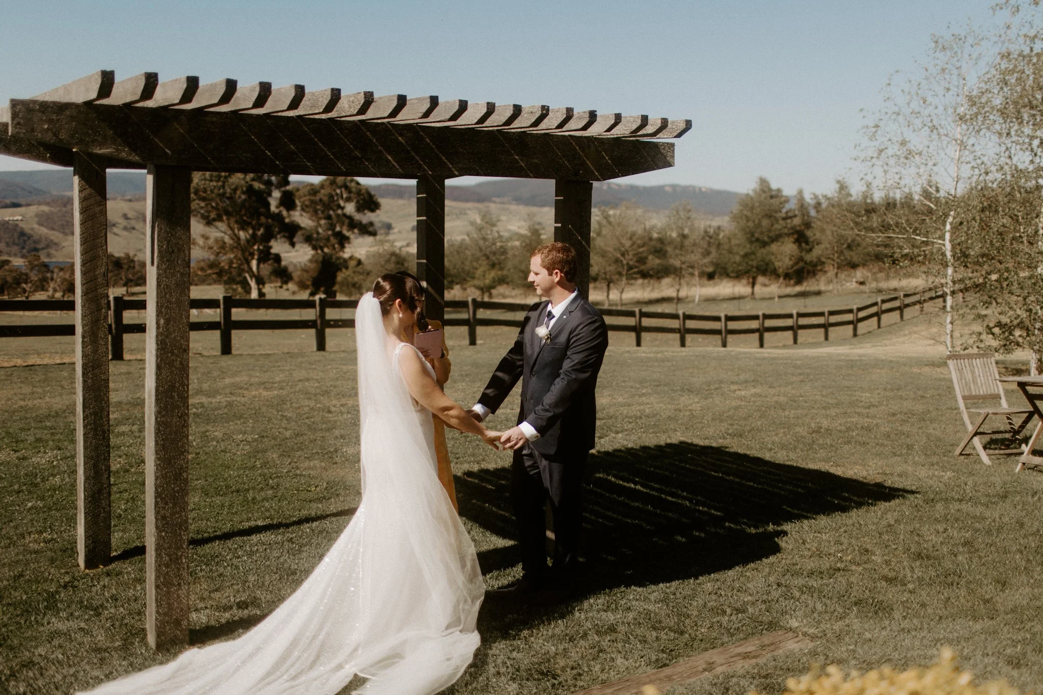 A wedding ceremony taking place outdoors with a bride and groom under a wooden arbor, in a scenic field with trees and mountains in the background at Seclusions Blue Mountains.