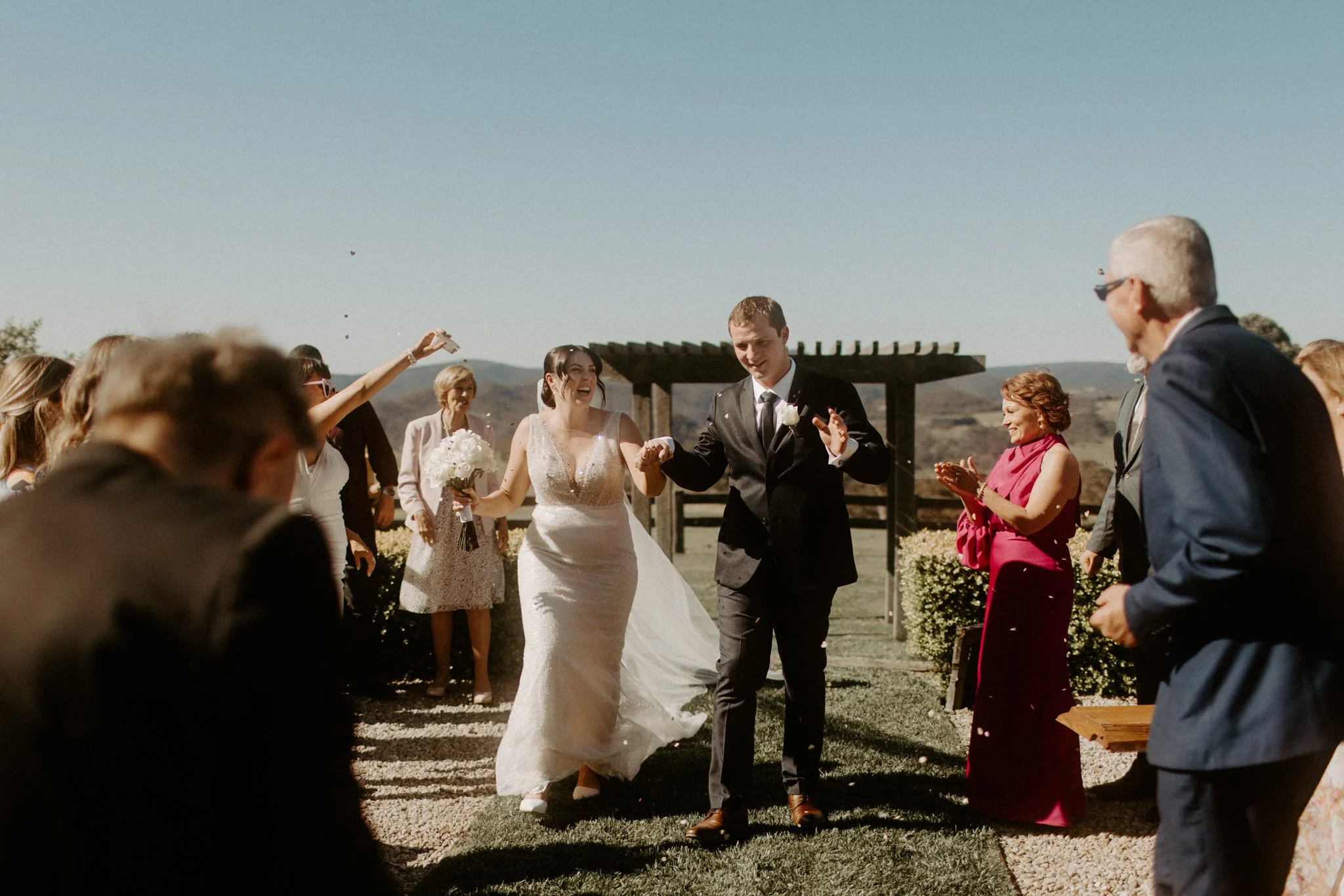 A bride and groom walking hand in hand, smiling, among friends and family during a Blue Mountains wedding at Seclusions Blue Mountains.