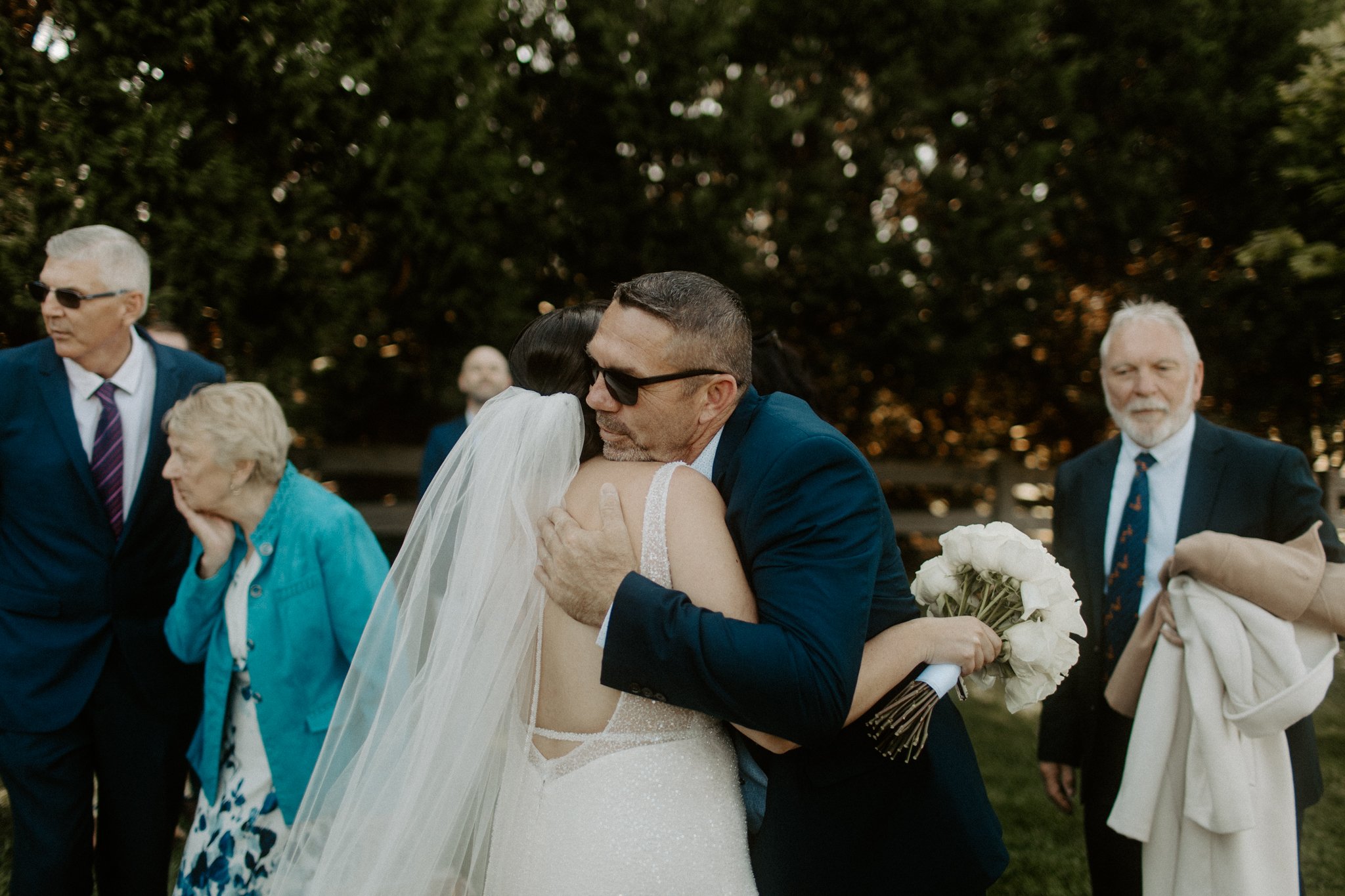 A couple hugging at a wedding, with the bride wearing a white gown and veil, holding a bouquet of white flowers, and the groom in a dark suit and sunglasses. Other guests are in the background outdoors.