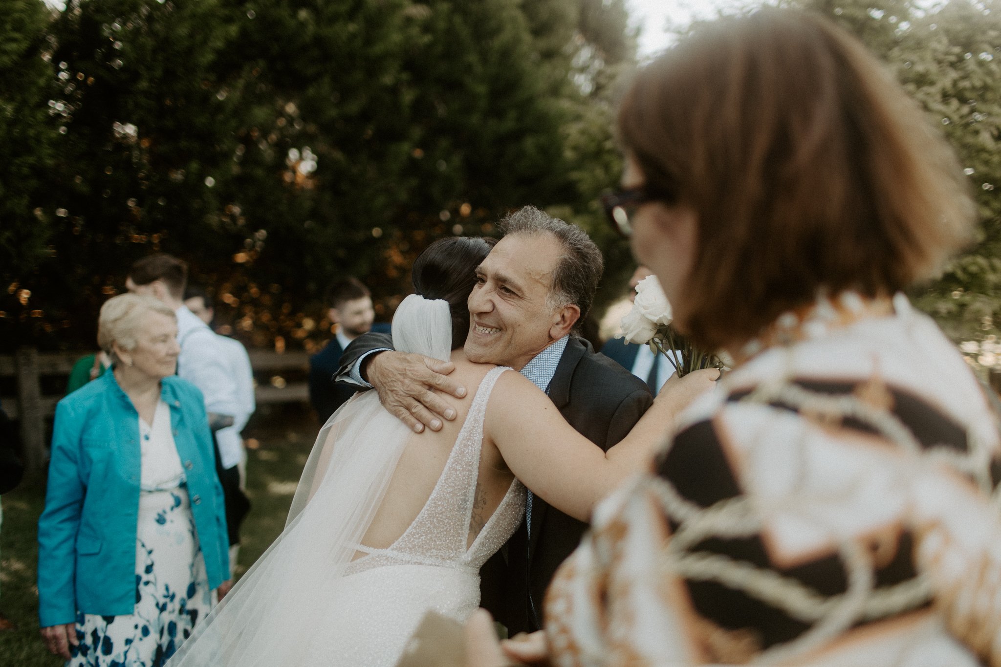 A bride hugging a smiling man outdoors at a wedding, with other guests in the background and a woman in the foreground.