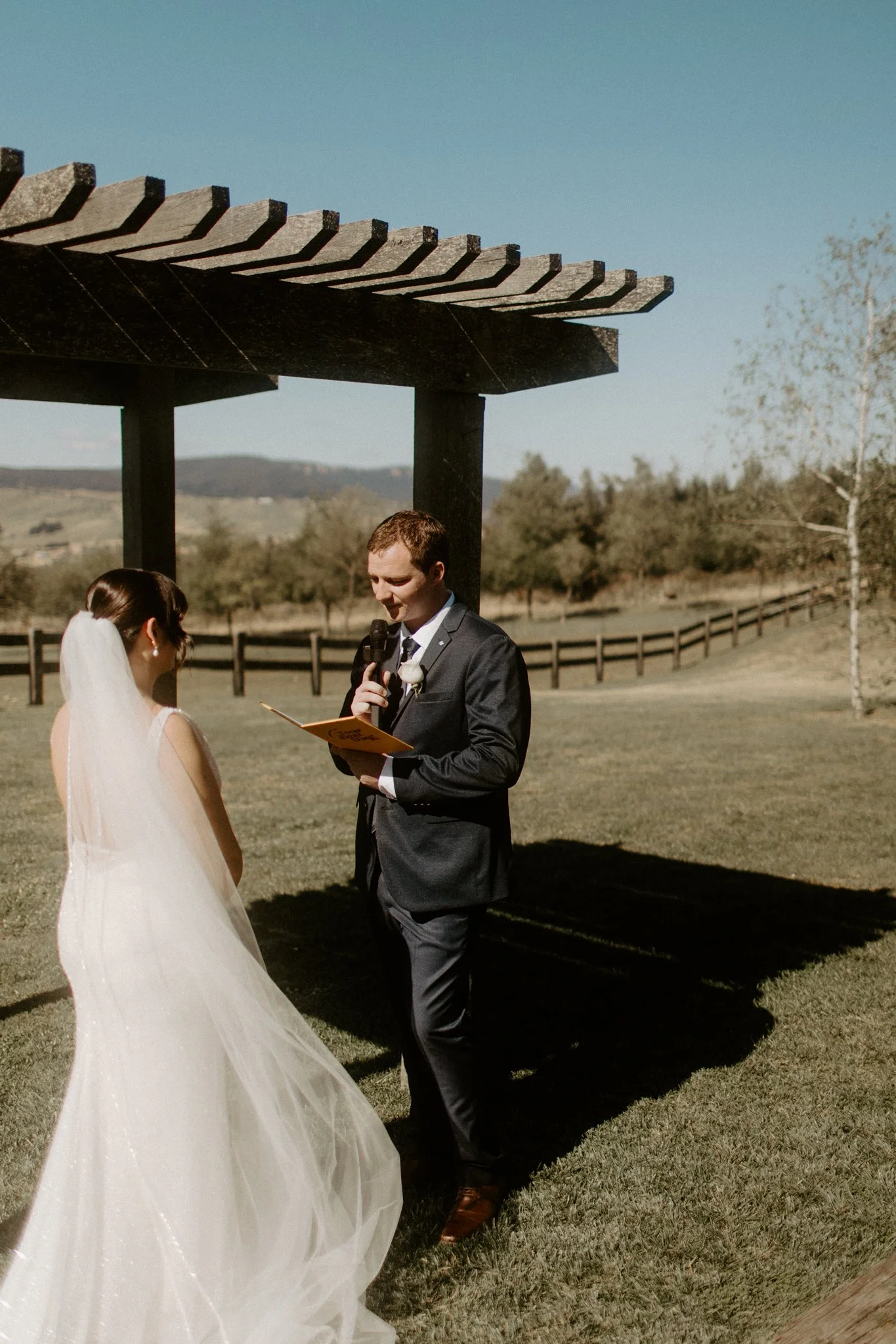 A wedding ceremony taking place outdoors with a bride and groom under a wooden arbor, in a scenic field with trees and mountains in the background at Seclusions Blue Mountains.
