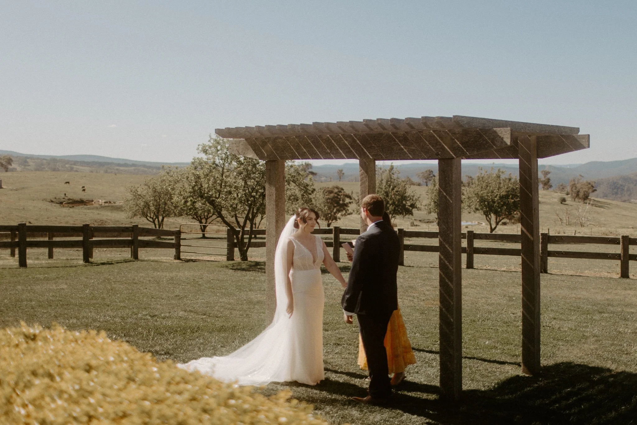 A wedding ceremony taking place outdoors with a bride and groom facing officiant under a wooden arbor, in a scenic field with trees and mountains in the background at Seclusions Blue Mountains.
