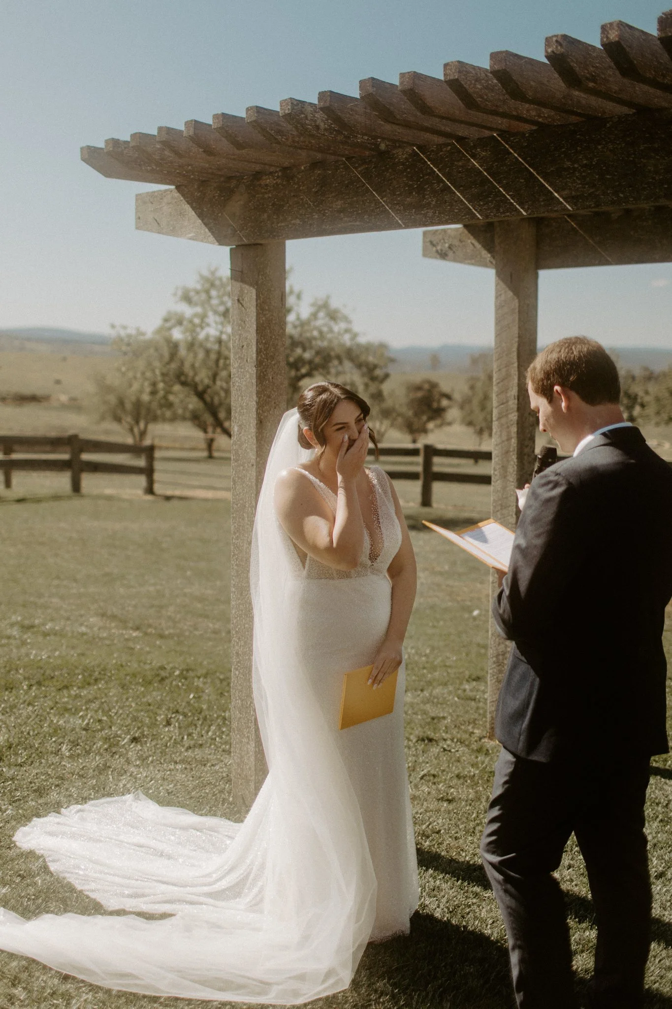 A wedding ceremony taking place outdoors with a bride and groom under a wooden arbor, in a scenic field with trees and mountains in the background at Seclusions Blue Mountains.