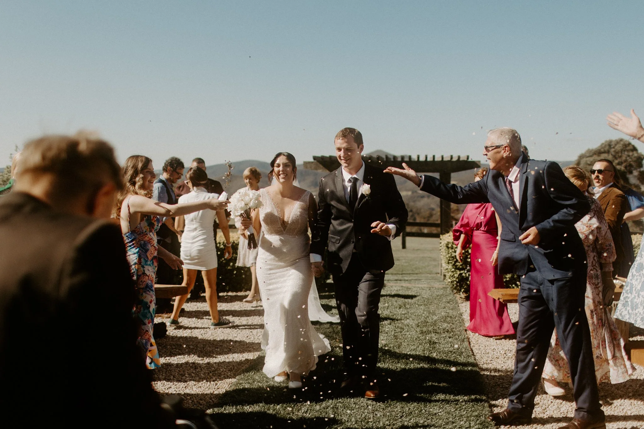 A bride and groom walking outdoors after wedding ceremony, surrounded by friends and family throwing flower petals, with a wooden structure and mountains in the background during a Blue Mountains wedding at Seclusions Blue Mountains.