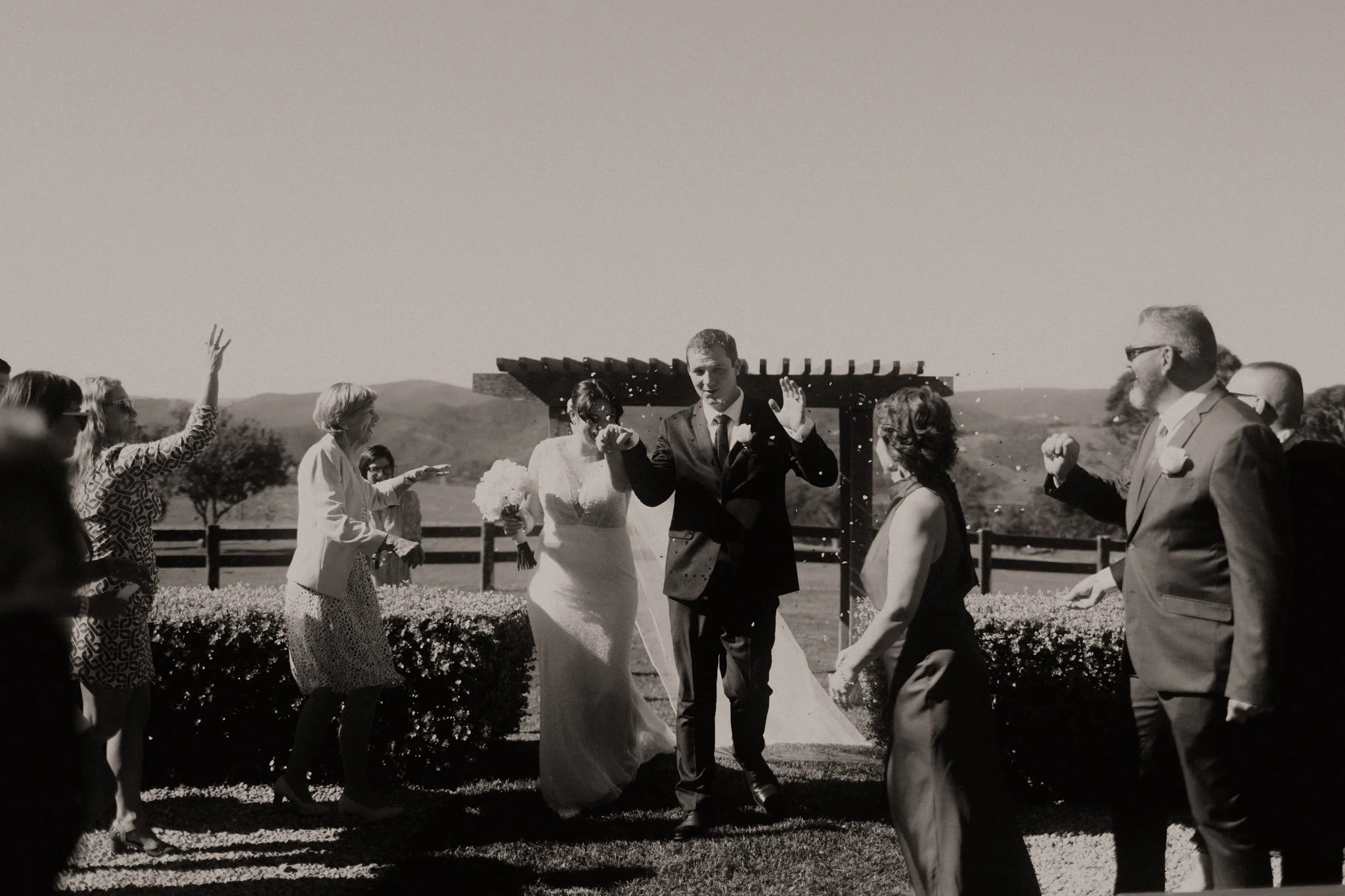 Black and white photo of a wedding celebration outdoors with guests throwing confetti as the bride and groom walk during a Blue Mountains wedding at Seclusions Blue Mountains.