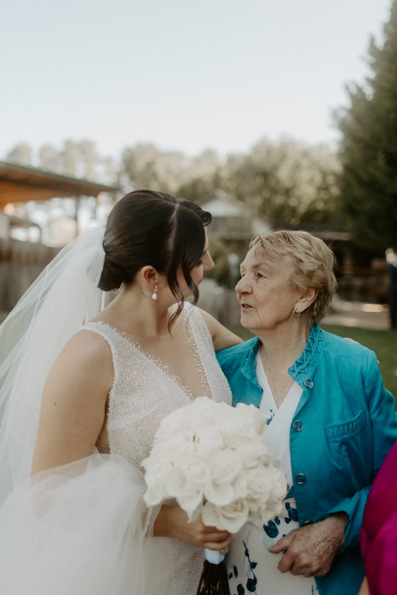 A bride in a wedding dress holding a bouquet of white roses is talking to an elderly woman outdoors during a wedding celebration. The bride has dark hair styled in an updo and is wearing pearl earrings.
