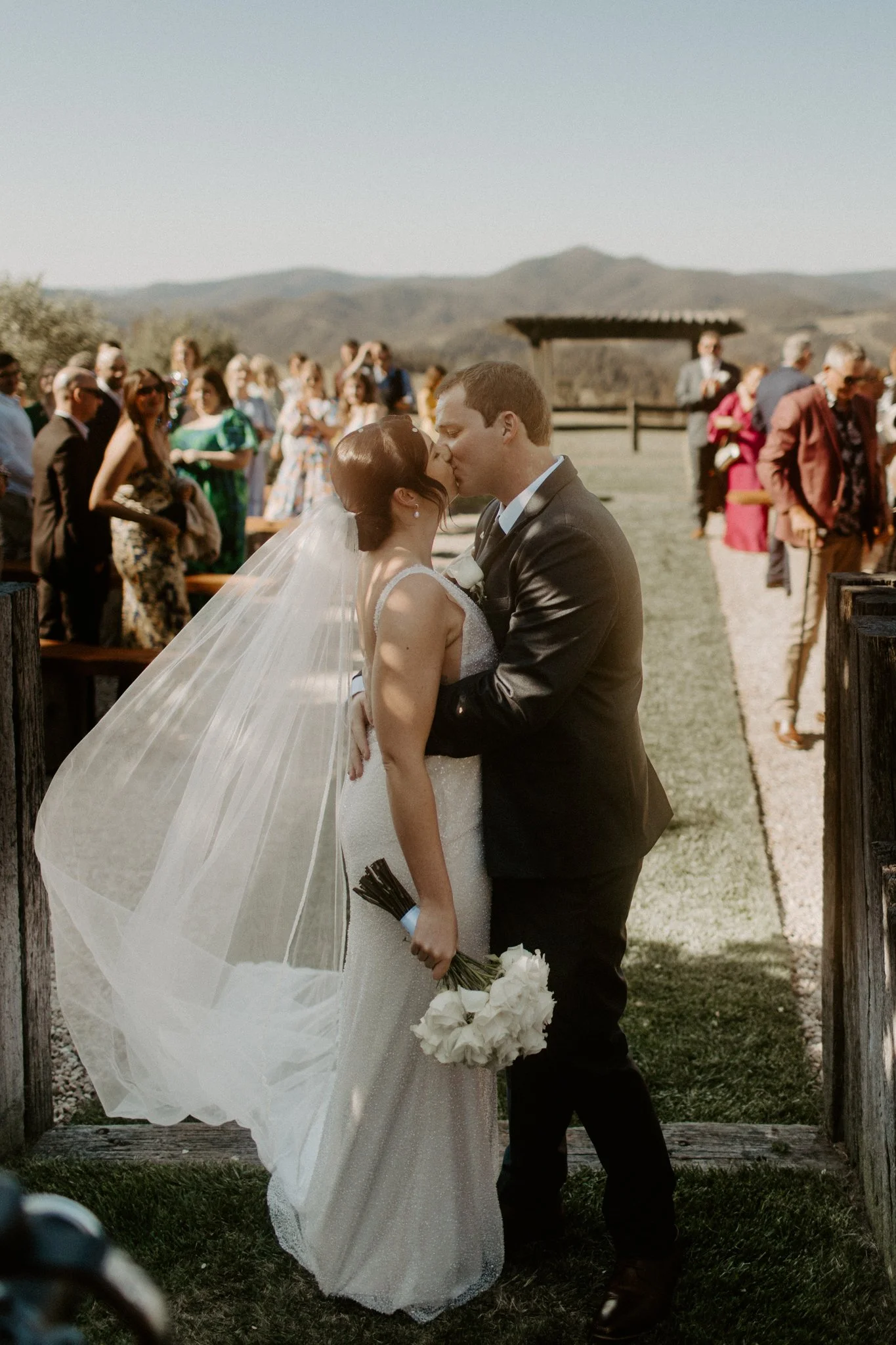 A bride and groom share a kiss at their outdoor wedding ceremony, surrounded by friends and family amidst a scenic landscape during a Blue Mountains wedding at Seclusions Blue Mountains.