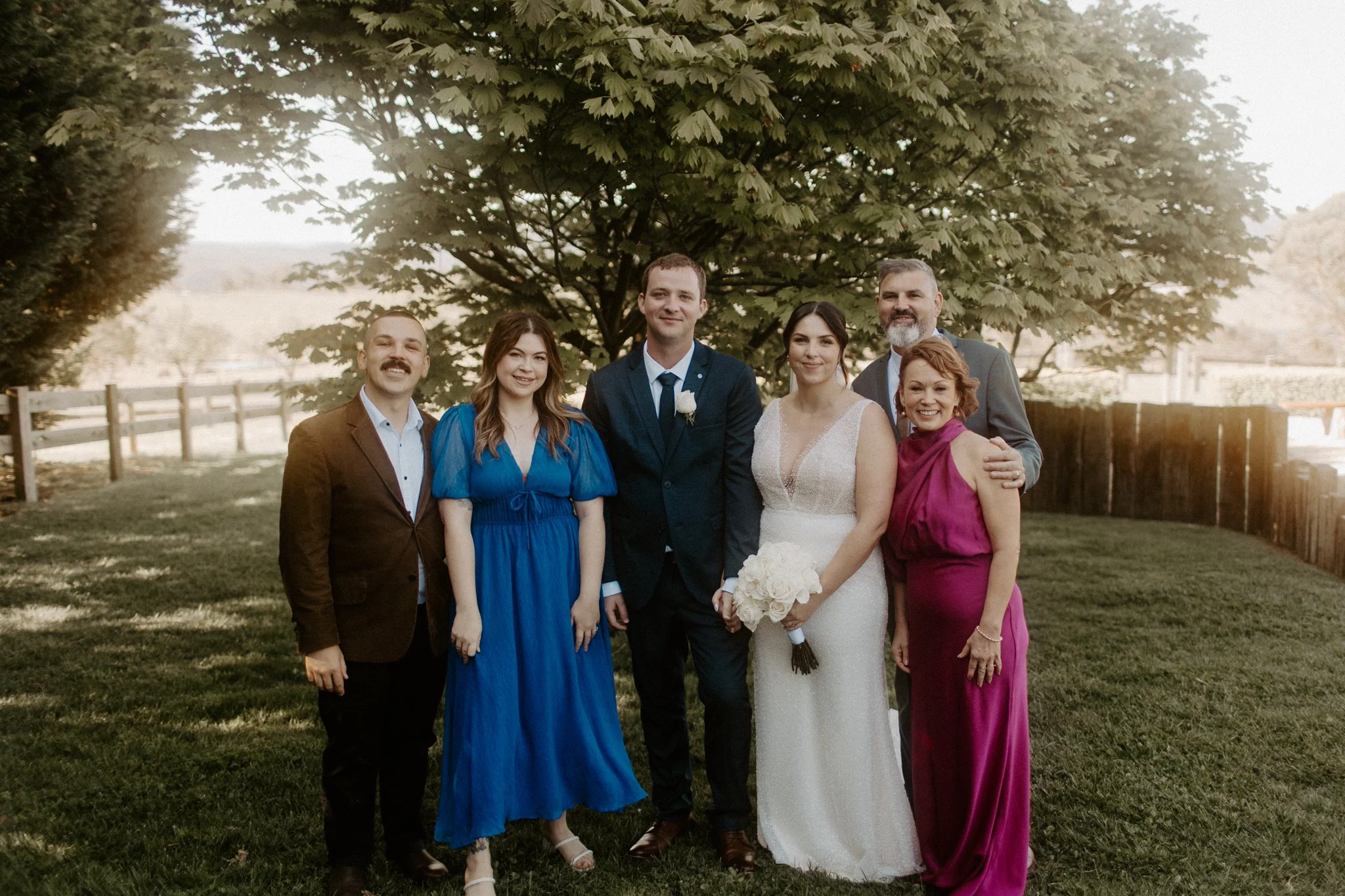 A group of six people, including a bride holding a bouquet, posing outdoors under a large tree on grass, with a wooden fence in the background. They are dressed in formal attire, likely at a wedding celebration.