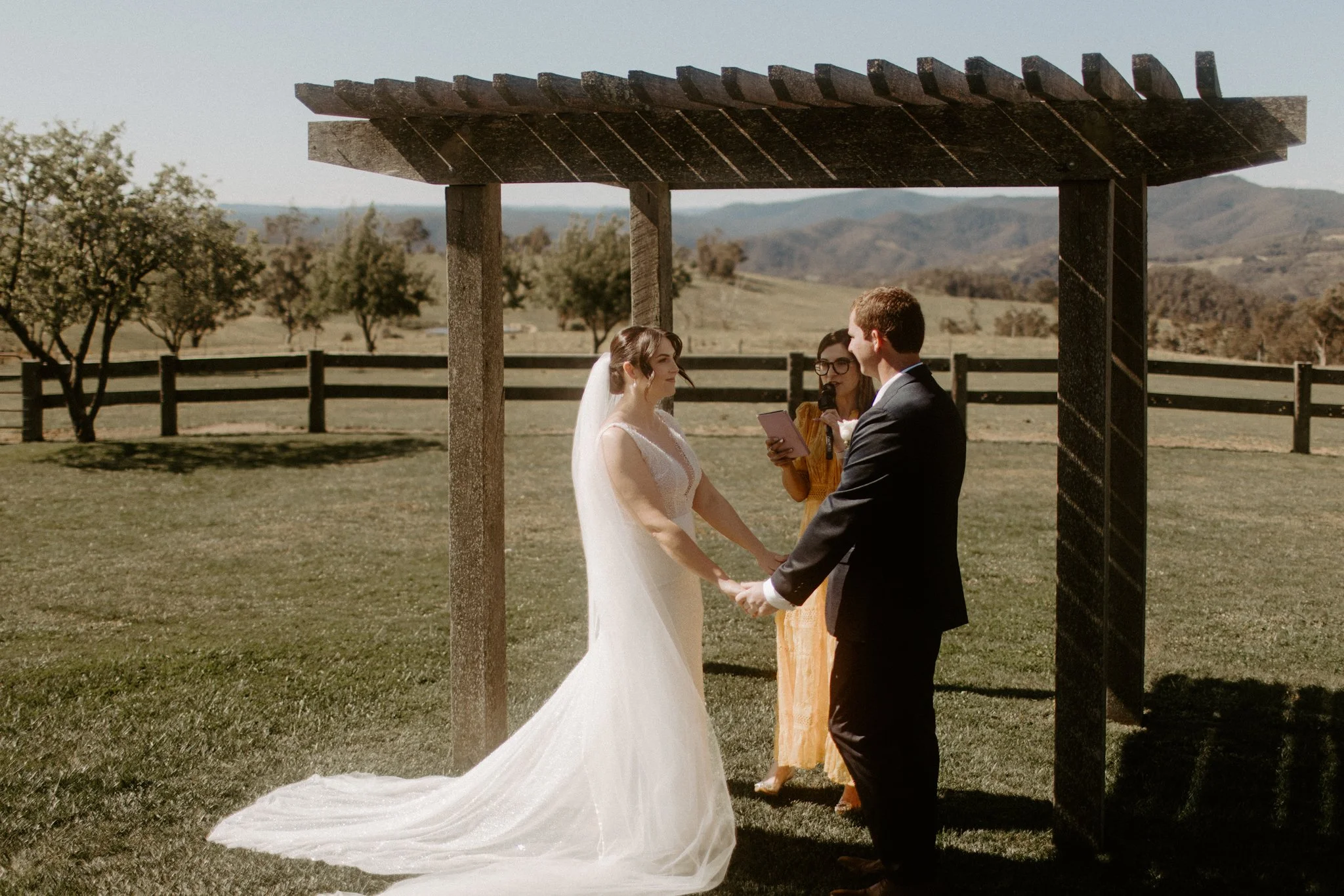 A wedding ceremony taking place outdoors with a bride and groom under a wooden arbor, in a scenic field with trees and mountains in the background at Seclusions Blue Mountains.