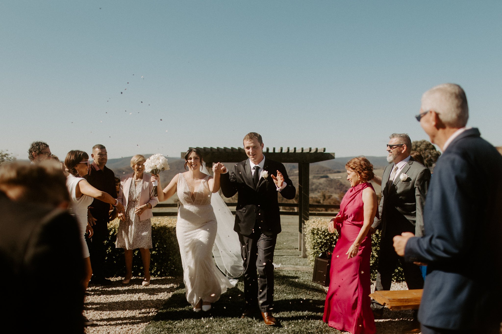 Bride and groom walking hand in hand down the aisle with guests on both sides celebrating under a clear blue sky during a Blue Mountains wedding at Seclusions Blue Mountains.