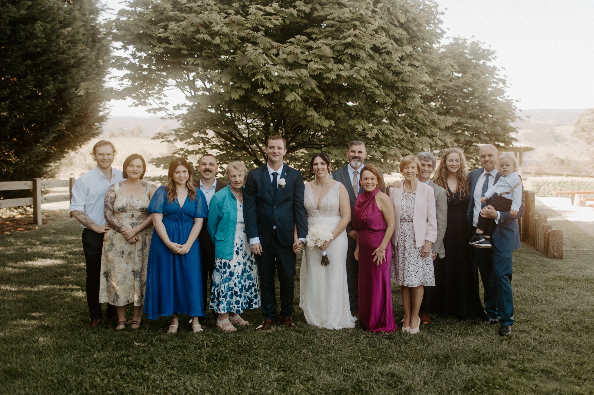Group of people, including a bride and groom, posing outdoors under a large tree at a wedding celebration.
