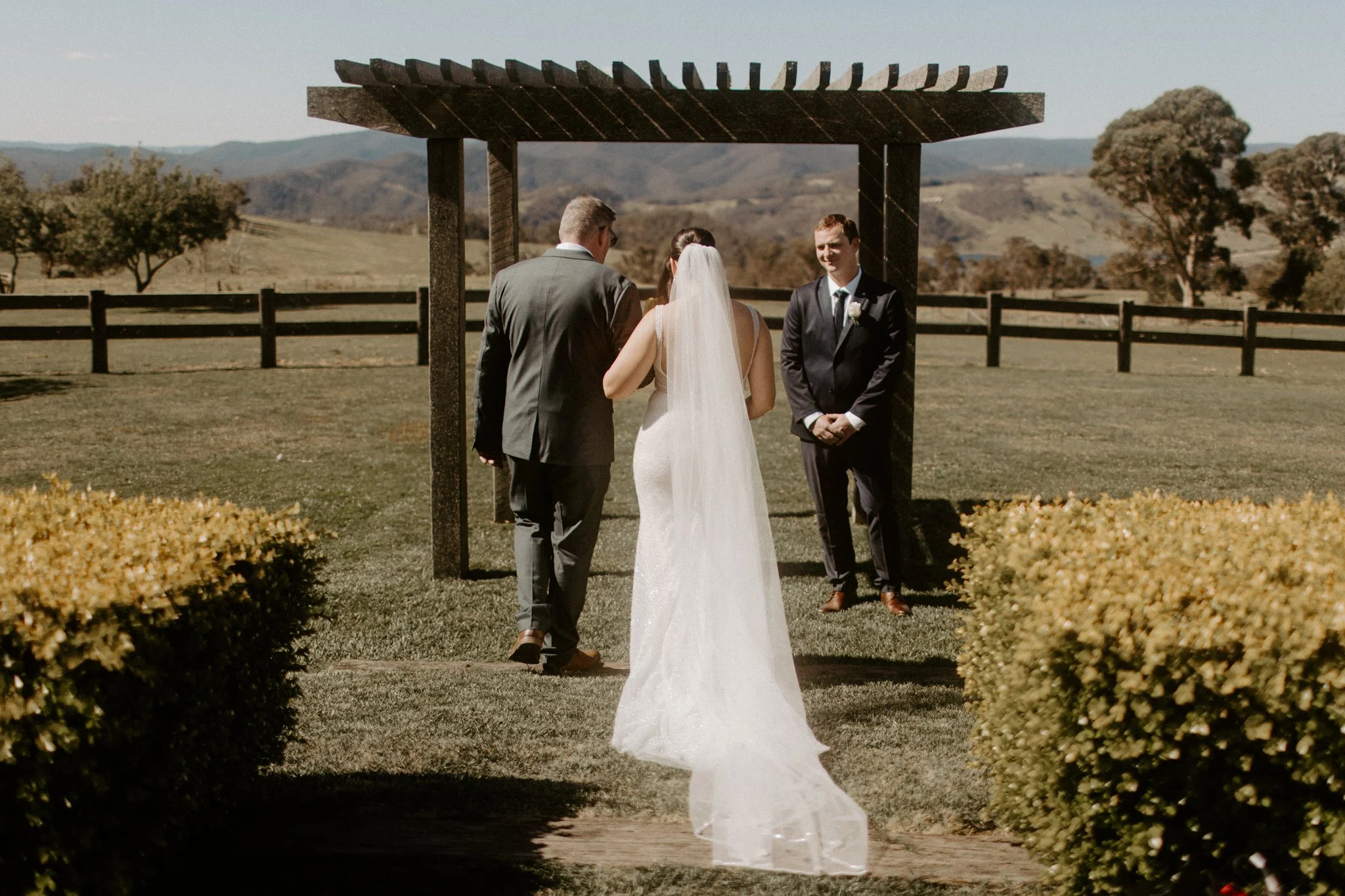 A wedding ceremony taking place outdoors with a bride and groom facing officiant under a wooden arbor, in a scenic field with trees and mountains in the background at Seclusions Blue Mountains.
