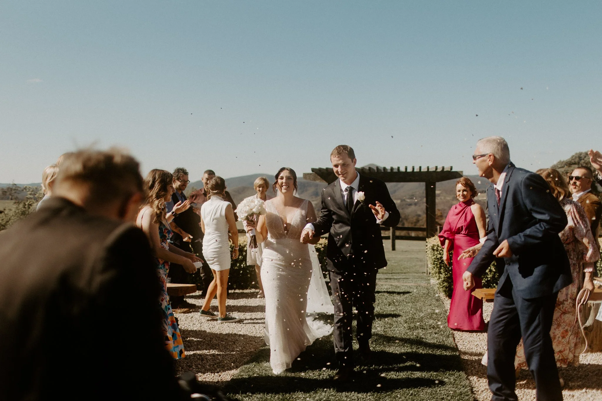 A wedding ceremony outdoors with a bride and groom walking down the aisle, guests on either side, and a clear blue sky during a Blue Mountains wedding at Seclusions Blue Mountains.