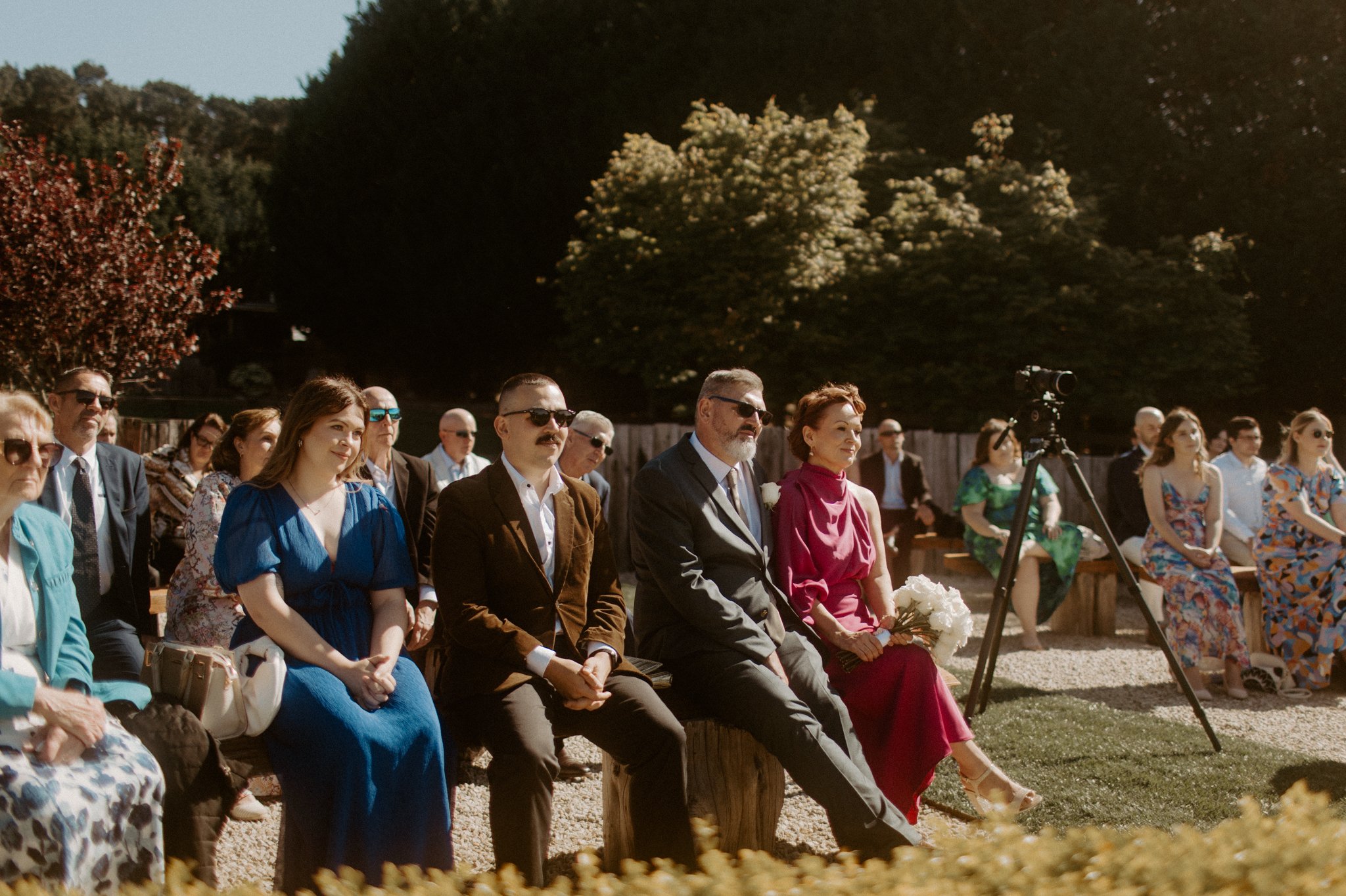 People attending an outdoor wedding ceremony, sitting on benches and chairs, with some wearing sunglasses and holding flowers, surrounded by trees and greenery at Seclusions Blue Mountains.
