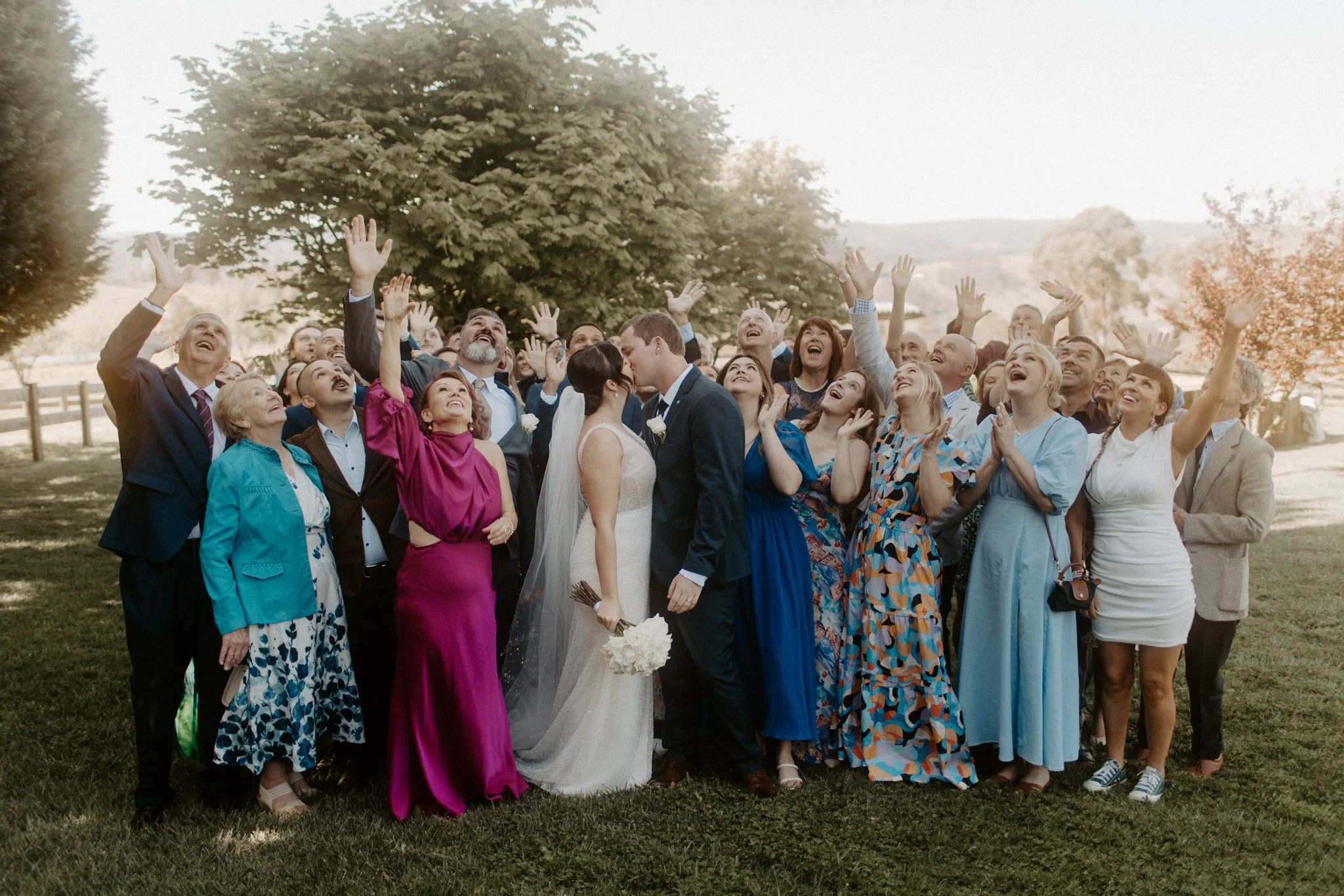Group of people at a wedding, including bride and groom, looking up and smiling in an outdoor setting with trees in the background.