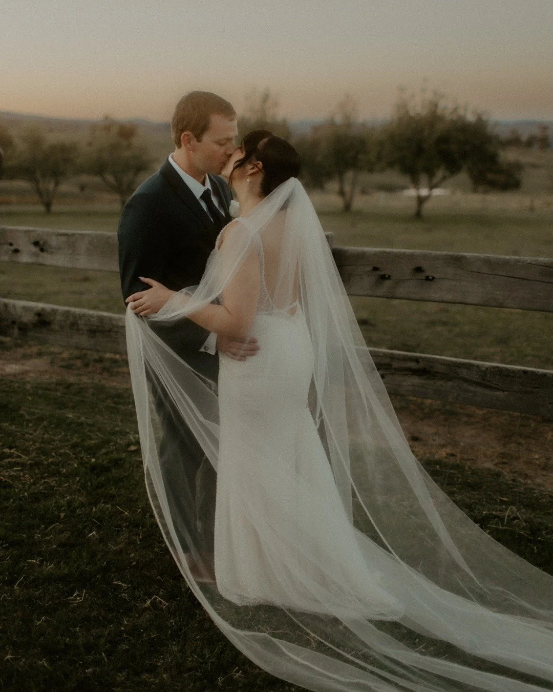 A bride and groom share a kiss outdoors at sunset, with the bride wearing a long veil and white gown, and the groom in a black suit.